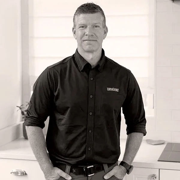 A man standing in a modern kitchen, wearing a black button-up shirt and a watch, with his hands in his pockets.