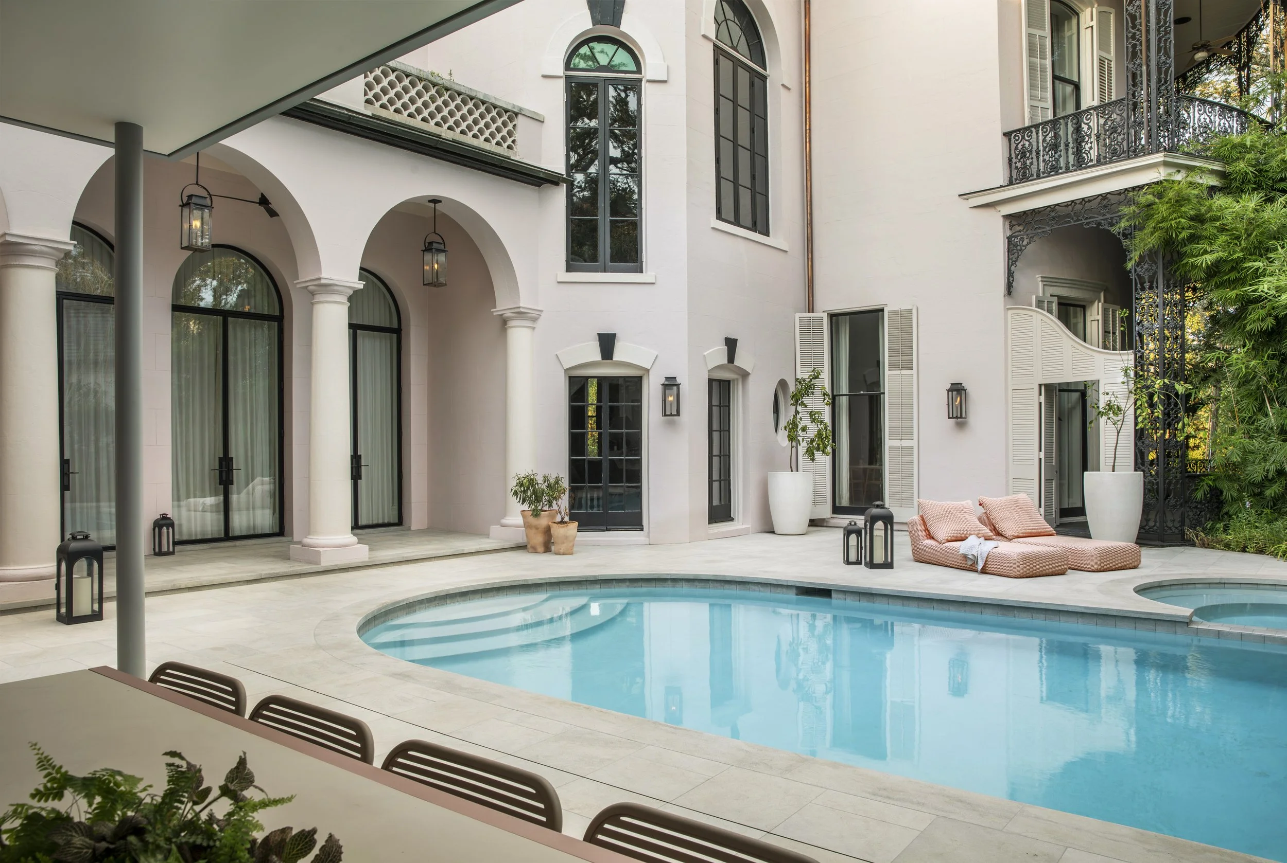 A luxurious outdoor pool area adjacent to a white house with arched windows, with lounge chairs, potted plants, and black lanterns decorated around the pool.