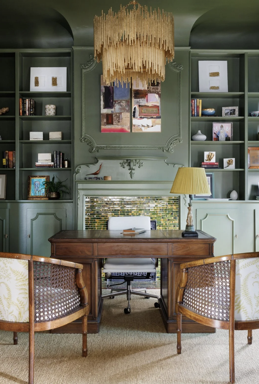 A home office with green built-in bookshelves, a wooden desk, a white office chair, a yellow table lamp, artwork, and decorative items.