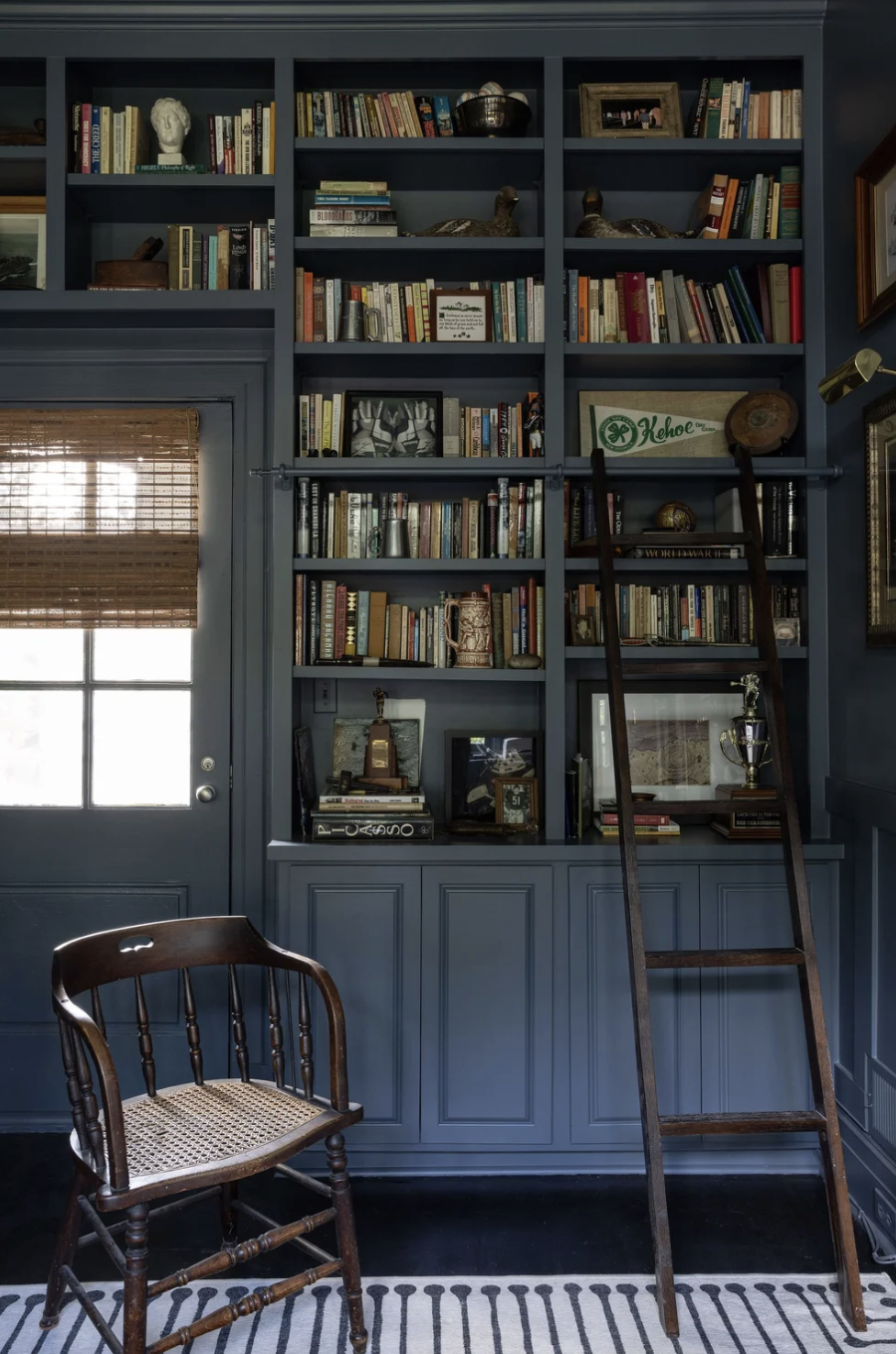 Blue built-in bookshelf filled with books, decorative objects, and framed photos, in a room with a window with bamboo blinds, a wooden chair with a woven seat, a ladder leaning against the bookshelf, and a striped rug.