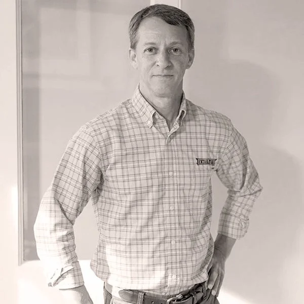 Man standing indoors, wearing a checkered button-up shirt, with one hand on his hip.