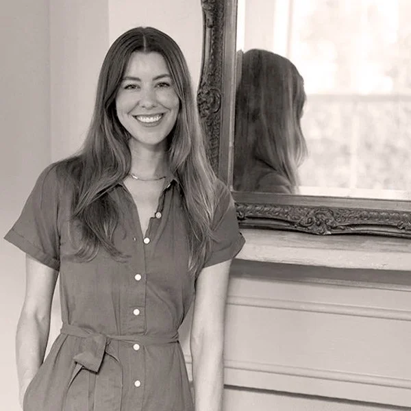A young woman with long wavy hair, smiling, standing next to a mirror, wearing a button-up dress with a waist tie.