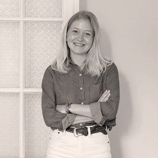 A woman with light-colored hair smiling, standing with her arms crossed in front of a patterned window in a room.