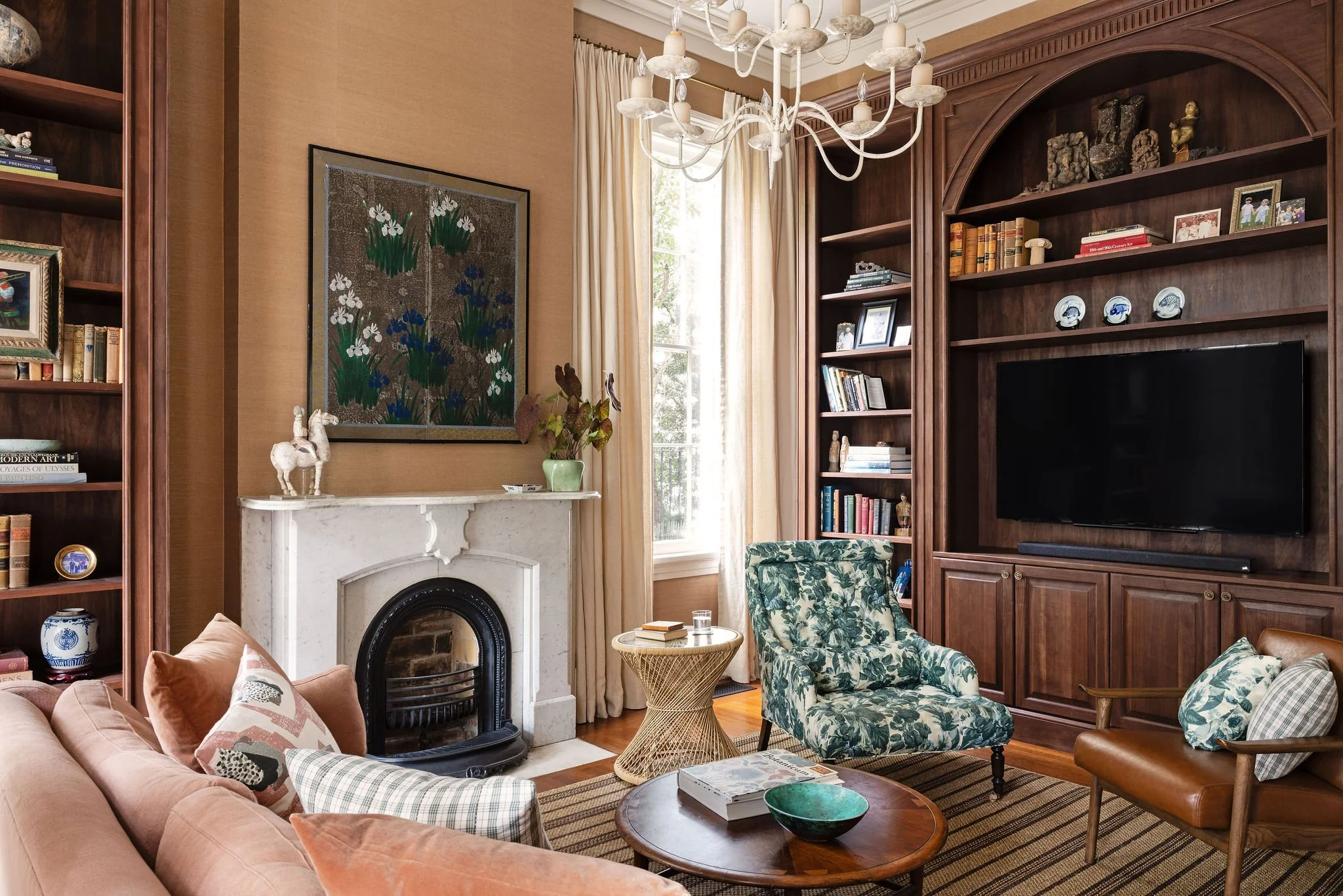 A cozy living room with a pink sofa, patterned armchair, and a marble fireplace featuring decorative sculptures and a framed artwork. There is a large wooden bookshelf, a chandelier, and a window with cream curtains letting in natural light.