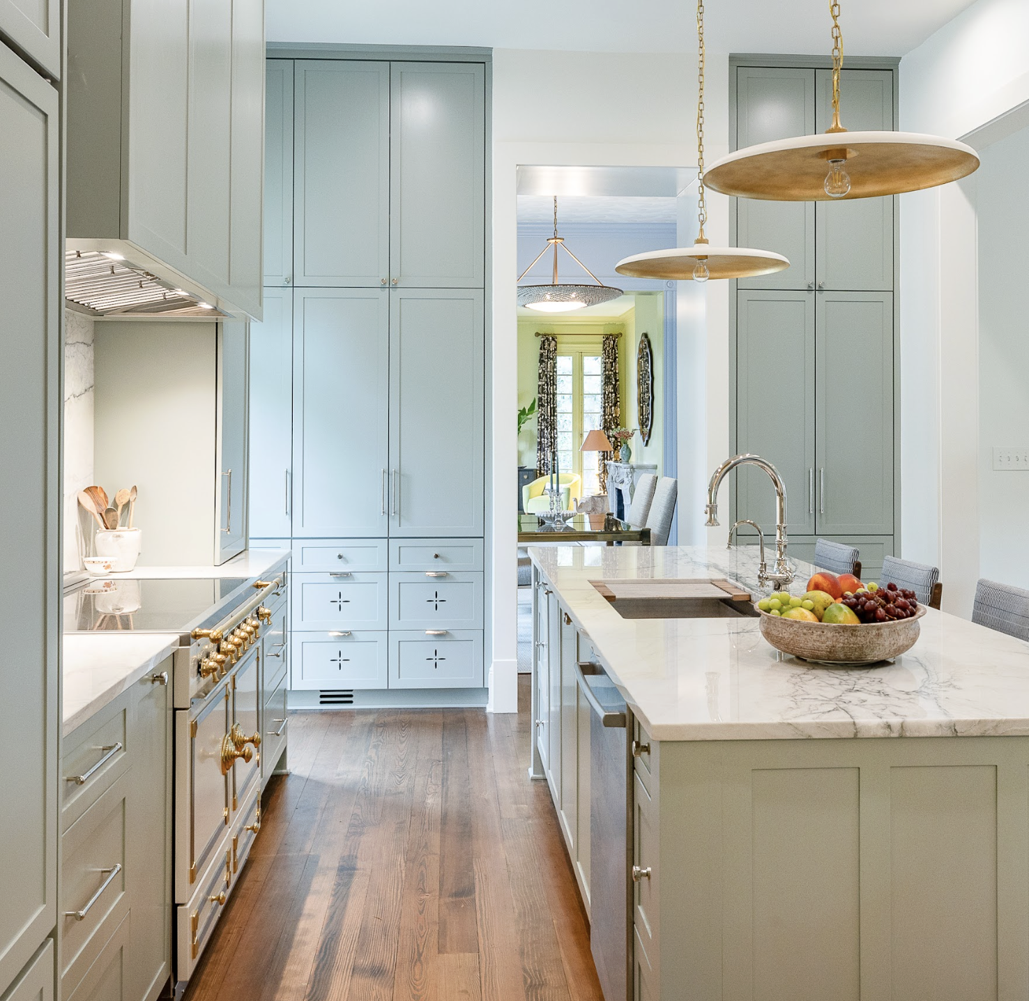 Modern kitchen with green cabinets, marble countertop island with a fruit bowl, stainless steel fixtures, and pendant lighting, opening to a dining area with a window and floral curtains.