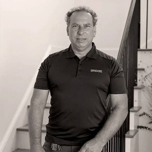 A man with curly hair wearing a black polo shirt standing on a staircase with a black railing in a house.