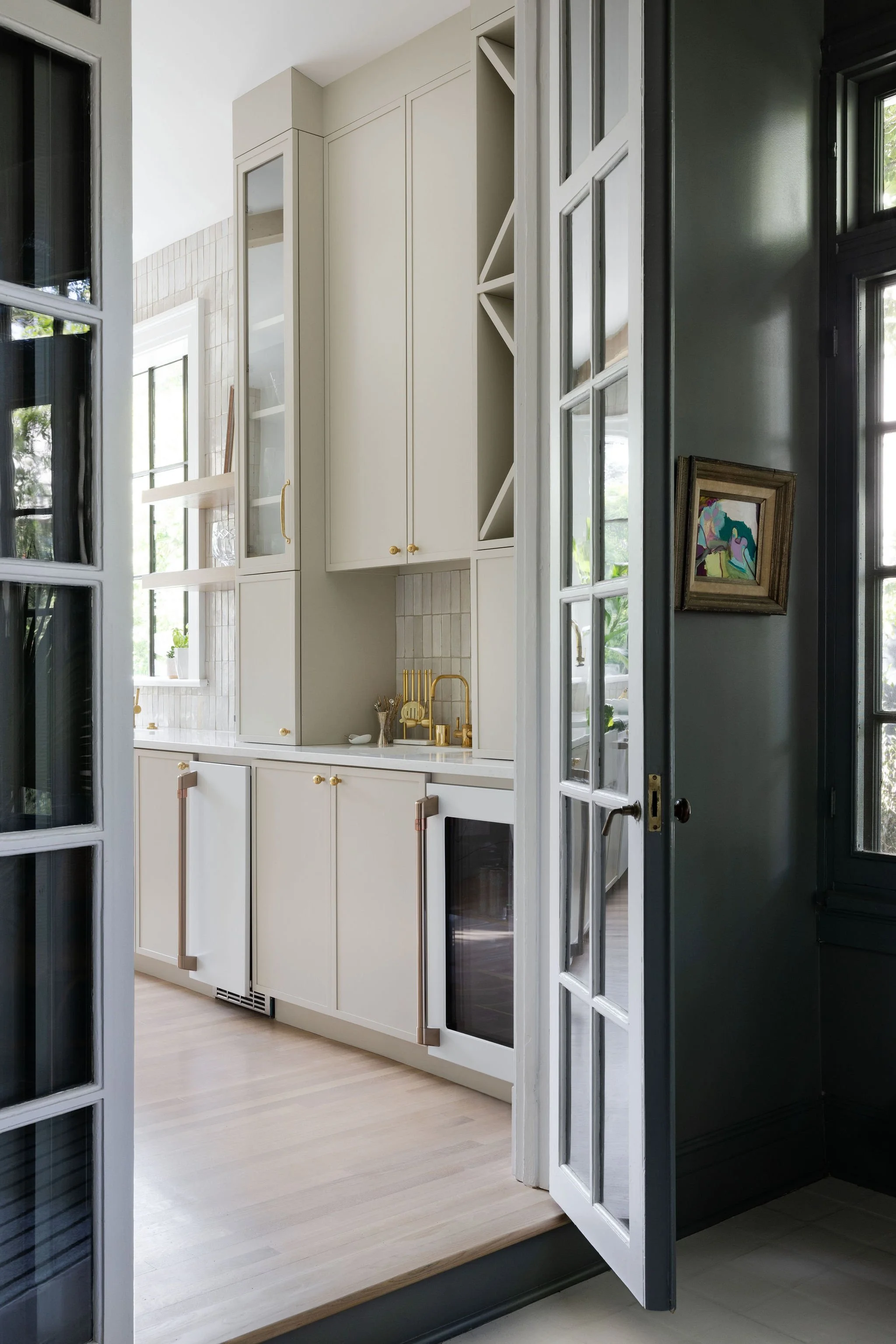 A kitchen viewed through open double glass doors with white frames, revealing white cabinets with gold hardware, a gold faucet, and a framed colorful artwork on the wall.
