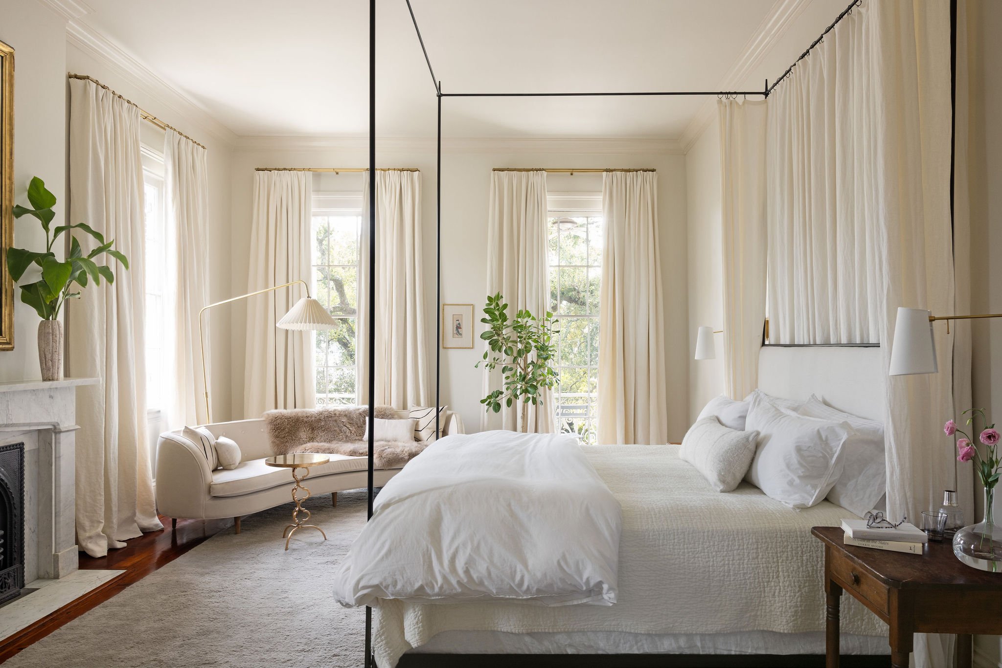 Bright bedroom with large windows, white curtains, a white bed with pillows, a wooden nightstand with books, a clear glass vase, and pink flowers, a white sofa with a fur throw, indoor plants, and a gold-framed mirror.