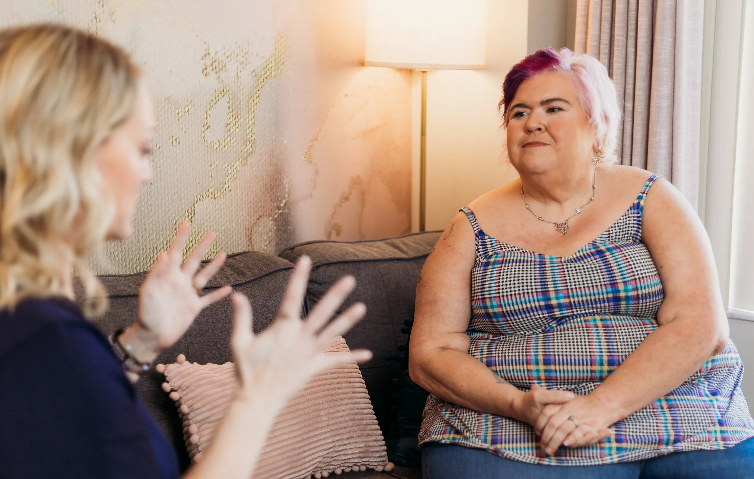 Two women talking in a living room, one with pink and white hair, wearing a checkered top, and the other with blonde hair, gesturing with hands.