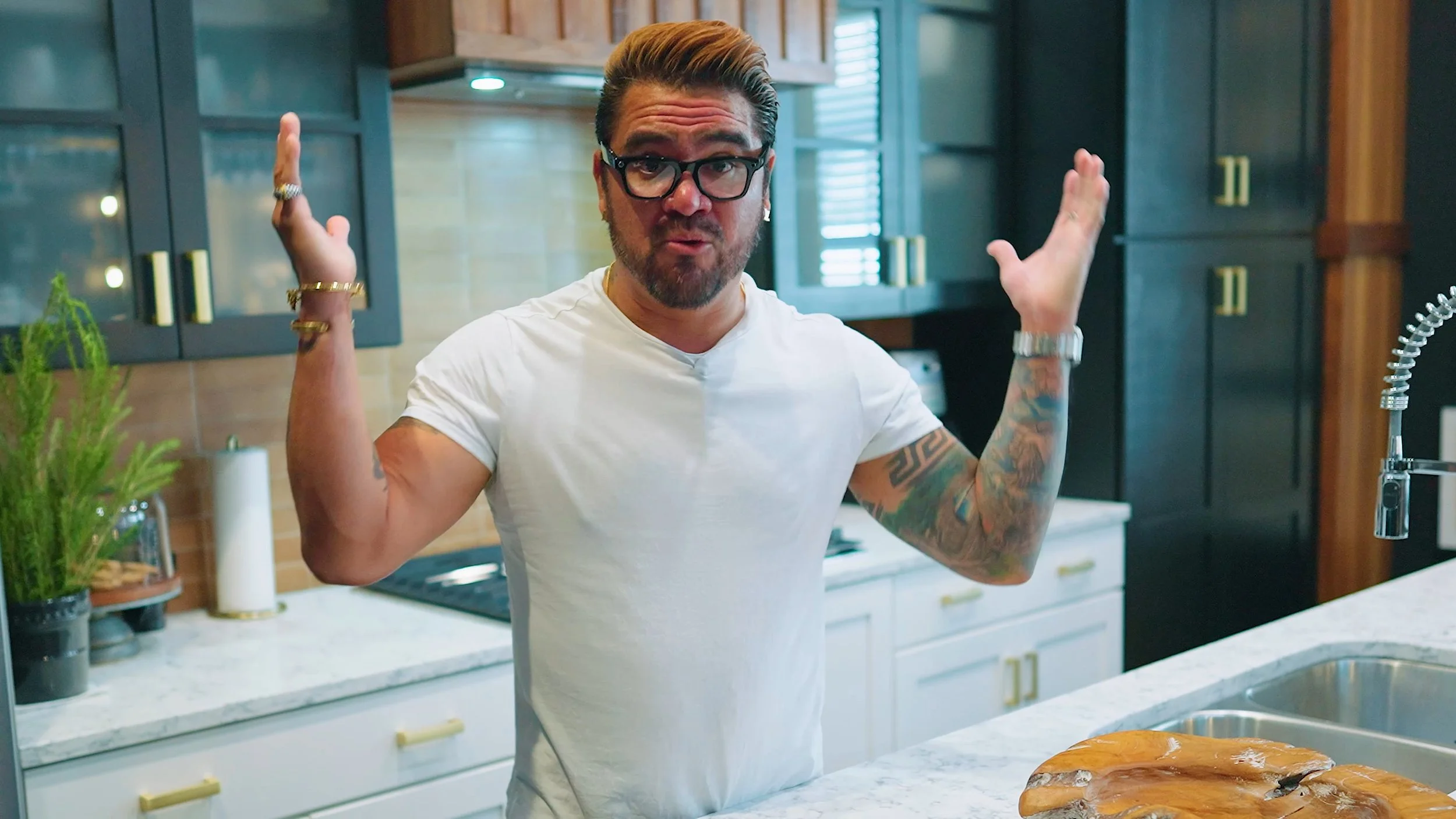 A man with styled hair, glasses, and tattoos on his arms, wearing a white t-shirt, standing in a modern kitchen with dark cabinets and a marble countertop, gesturing with his hands.