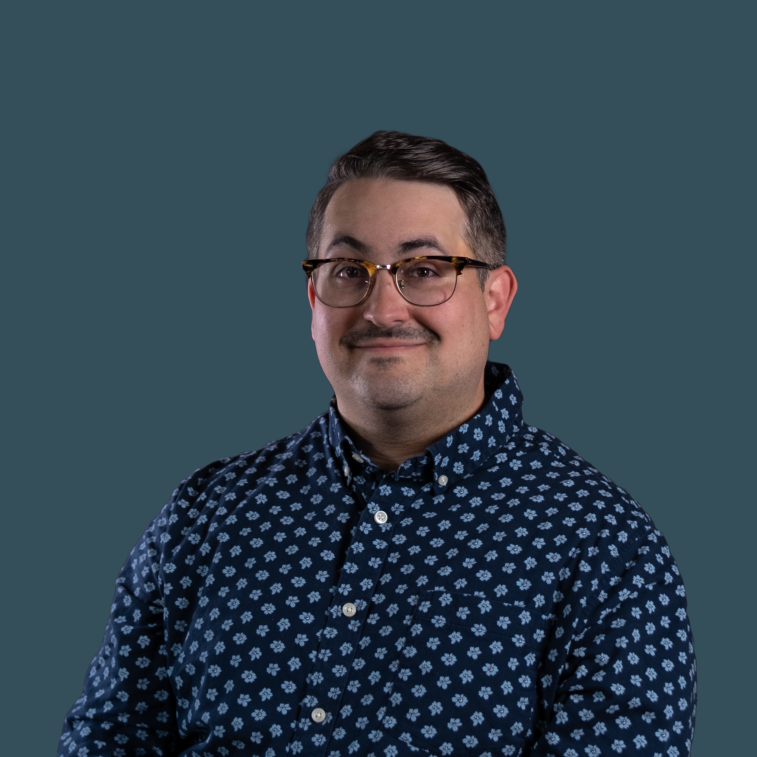 Close-up of a man with dark hair, glasses, and a patterned shirt, smiling against a plain teal background.