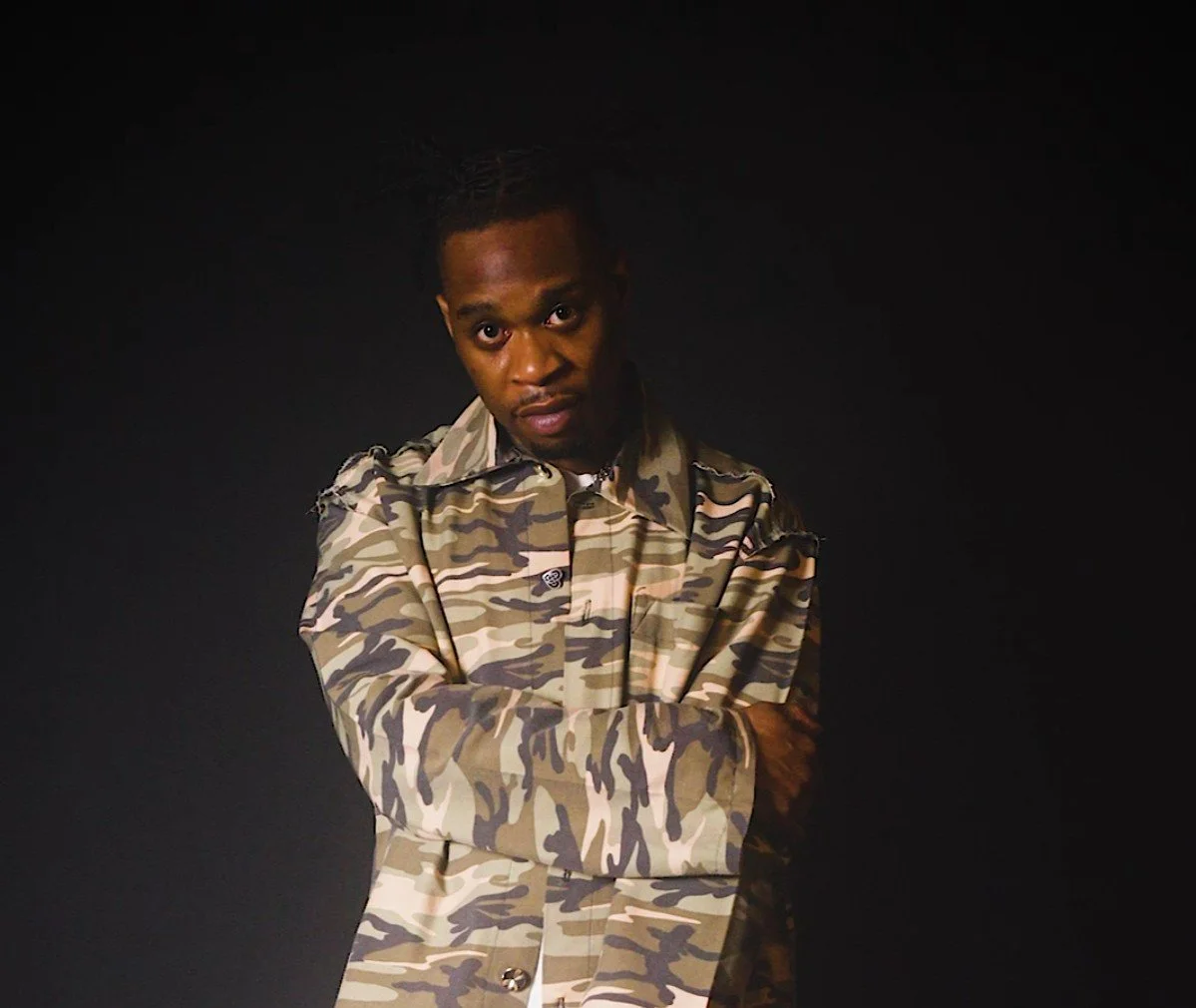 A young man with short hair wearing a camouflage jacket, standing against a black background, looking directly at the camera with crossed arms.