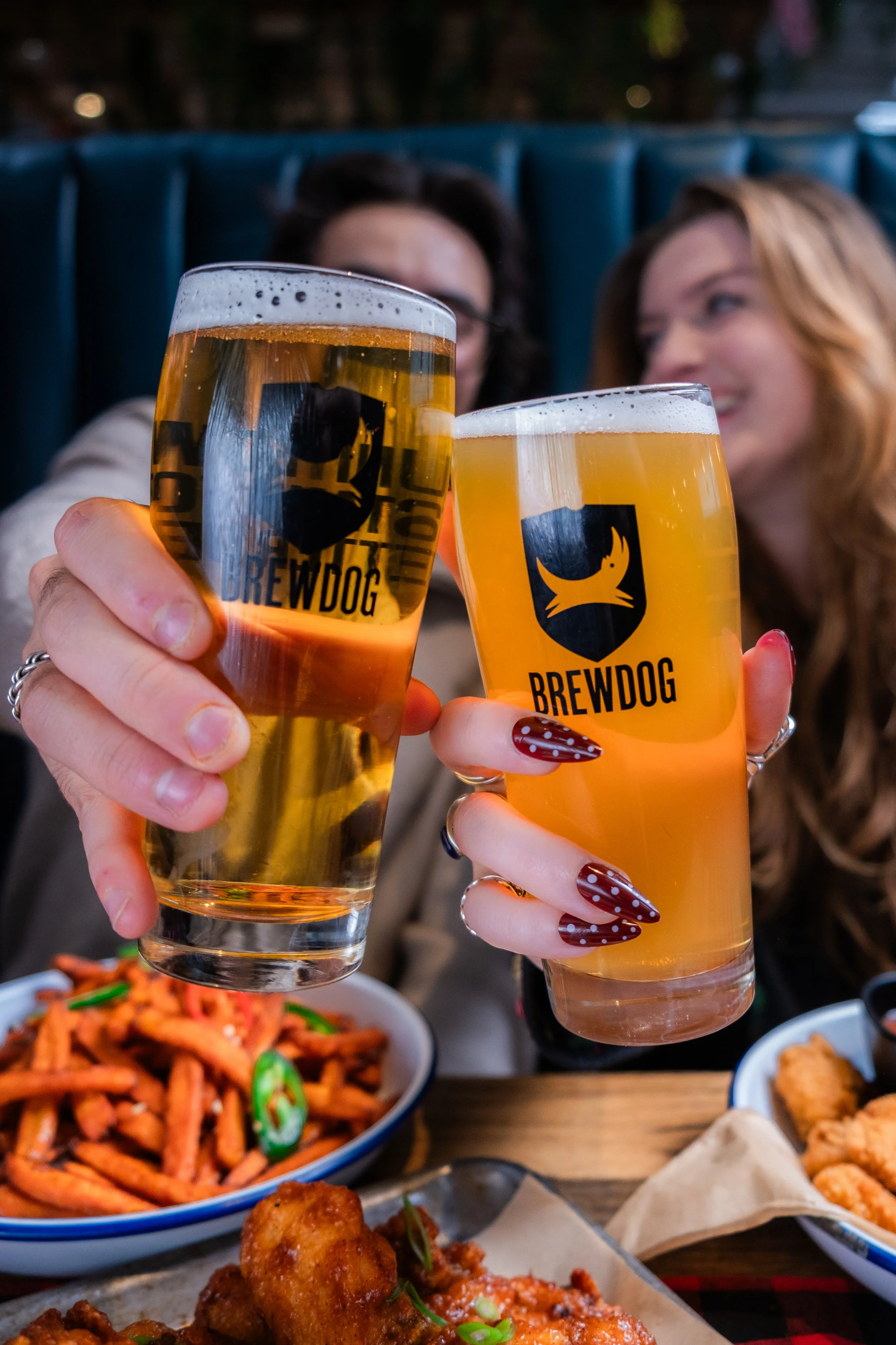 Two women toasting with Brewdog branded beer glasses at a restaurant, with plates of food on the table. The food includes fried chicken, snacks, and a colorful noodle dish.