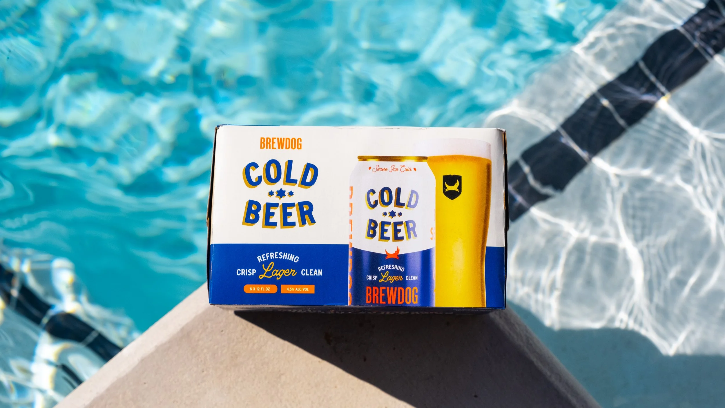 A box of Brewdog Cold Beer near a swimming pool with clear blue water, pool steps, and a concrete edge.