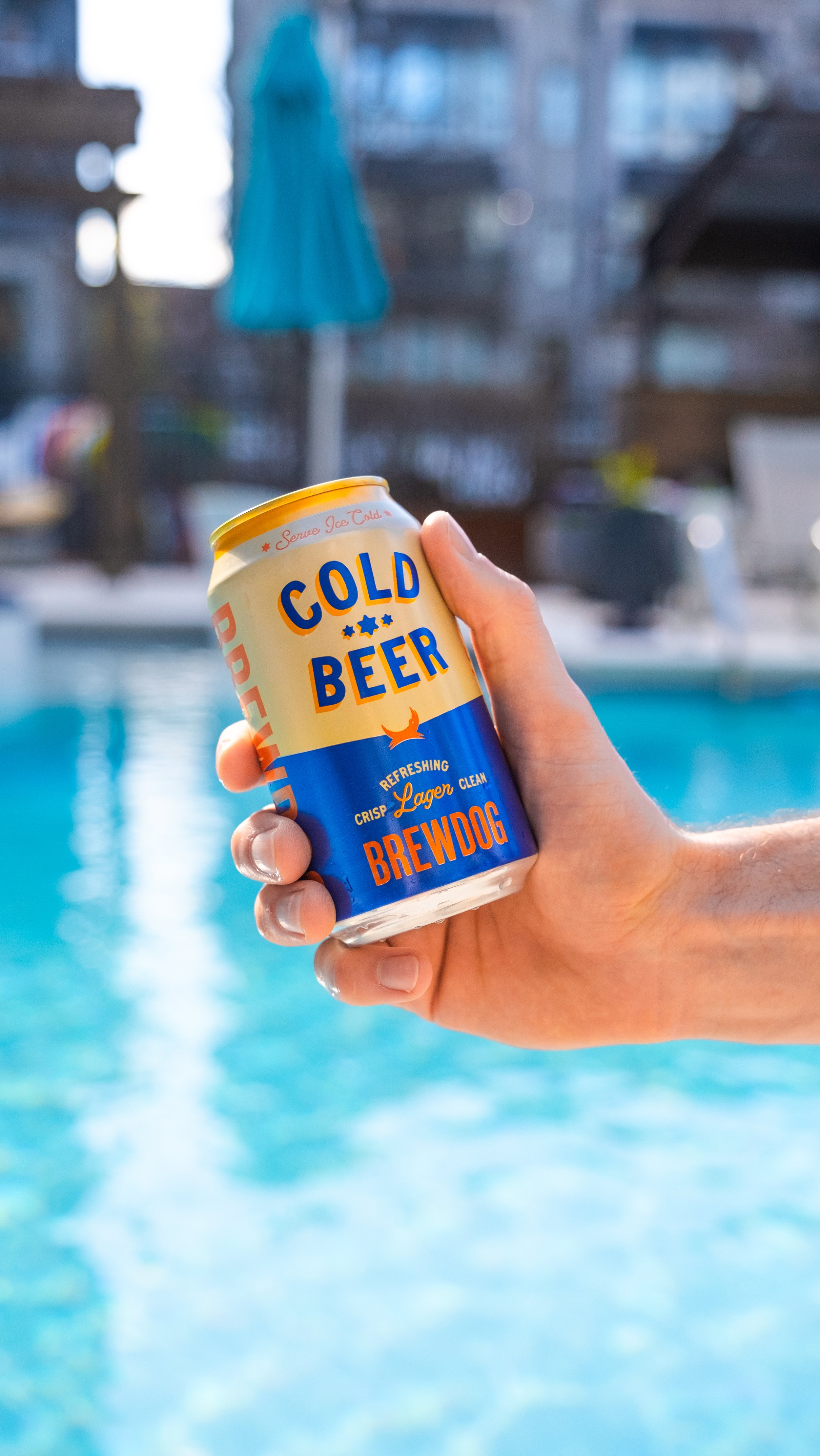 A hand holding a can of cold beer with a pool in the background and an outdoor living space with a blue umbrella.