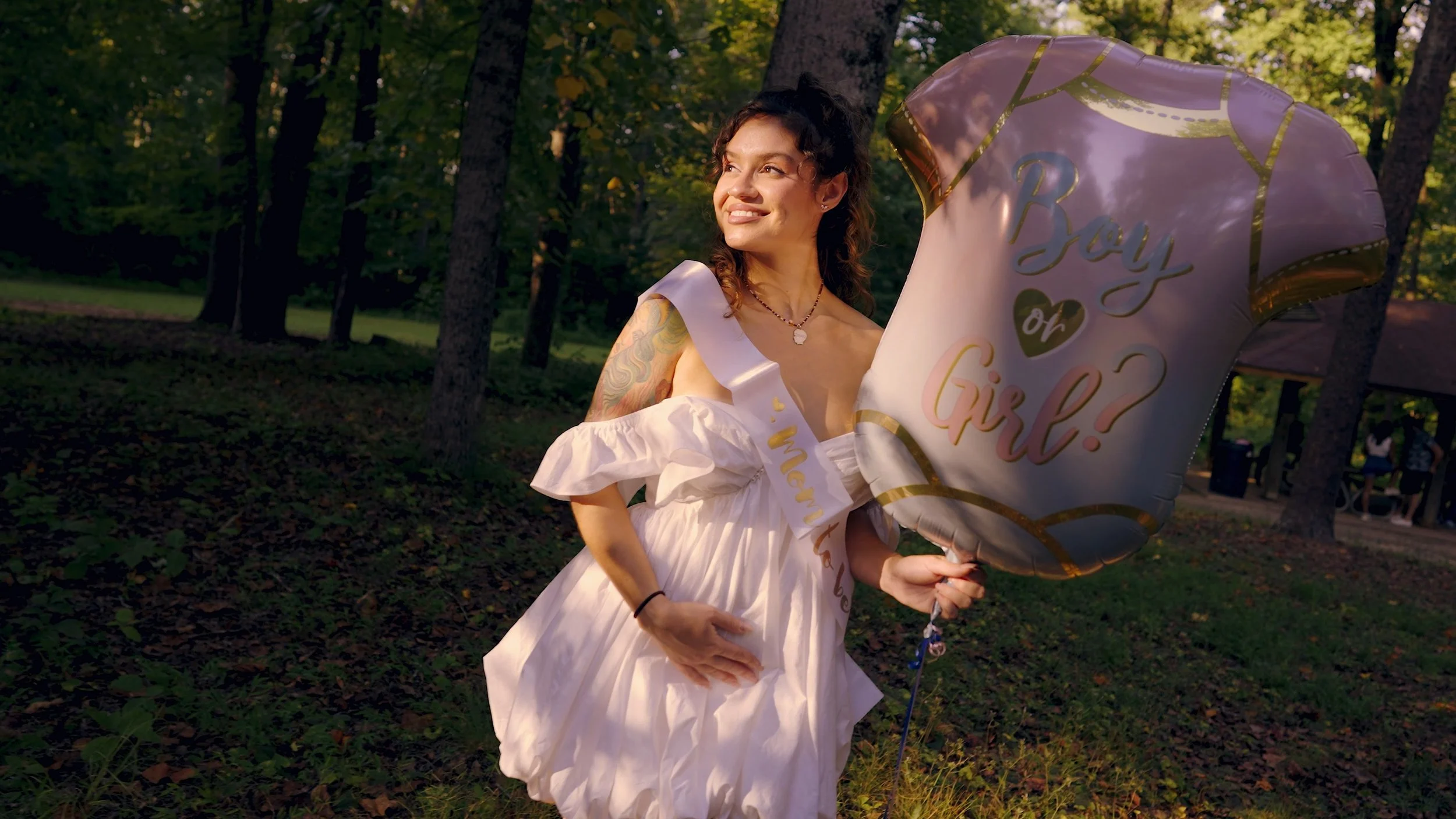 A young woman in a white dress with a sash and a necklace, smiling and holding a balloon that reads 'You or Girl?' in a wooded park area.