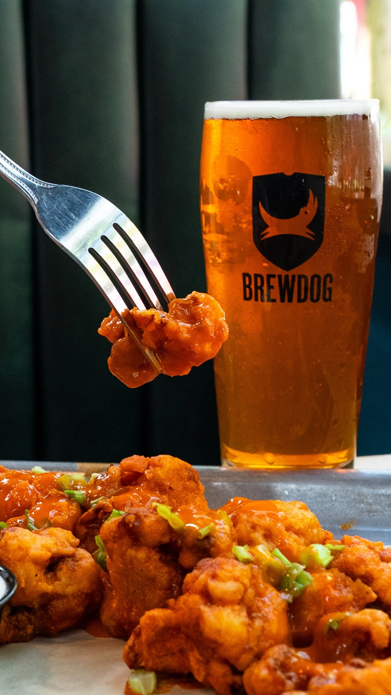 A glass of amber beer with a logo labeled "BREWDOG" next to a plate of spicy fried chicken coated in sauce with green onion garnishes.