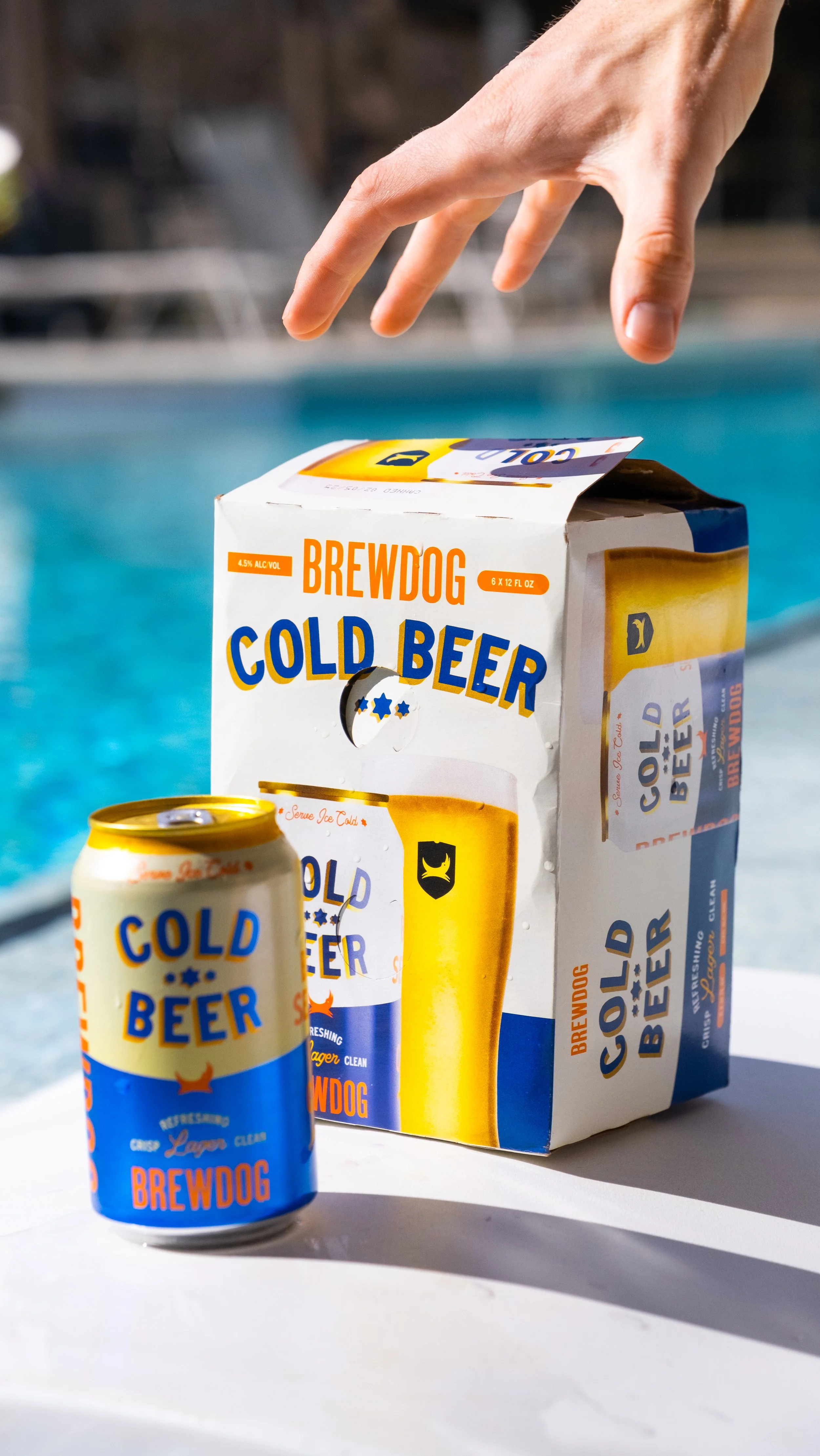 A box and a can of Brewdog Cold Beer, placed on a white surface near a pool with blue water. The box is white with orange, blue, and yellow labels, and the can has similar colorful branding.