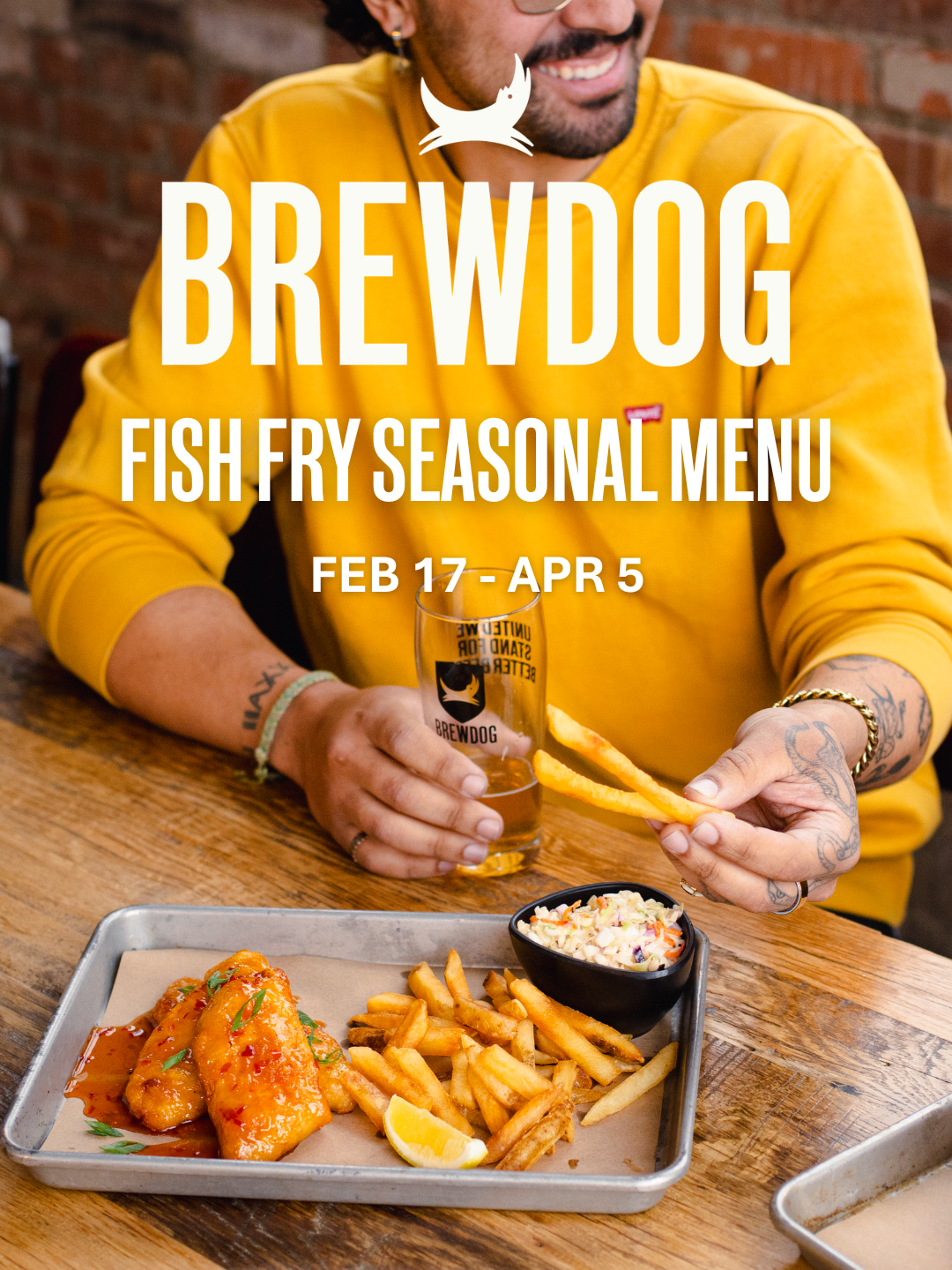 A person wearing a yellow sweatshirt is holding a glass of beer with the Brewdog logo, sitting at a wooden table with a tray of fish fry, French fries, and coleslaw.