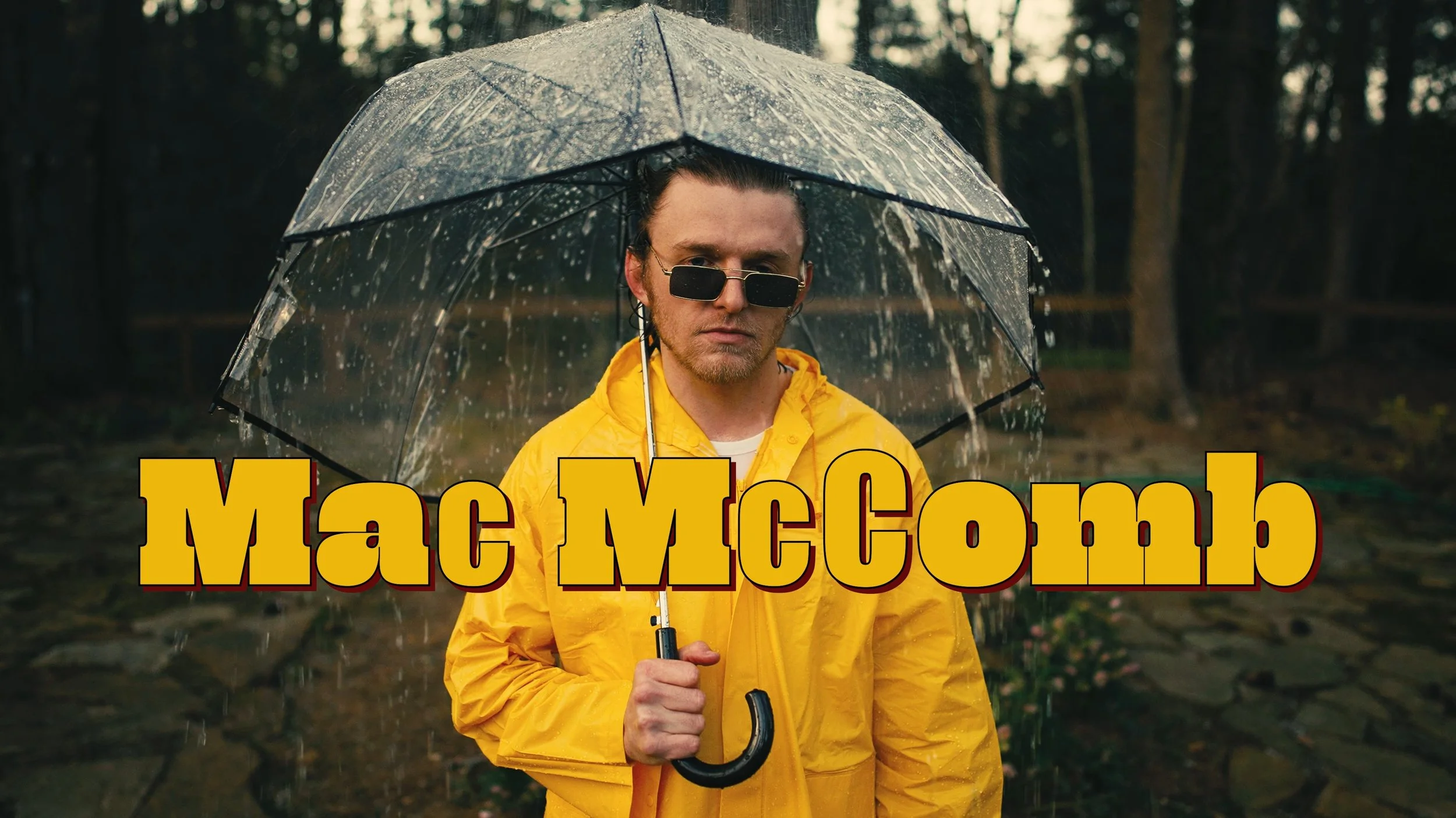 A man in a yellow rain jacket holding an umbrella stands outdoors in the rain, with trees in the background. Overlaid on the image are the words 'Mac McComb' in large, yellow and red text.
