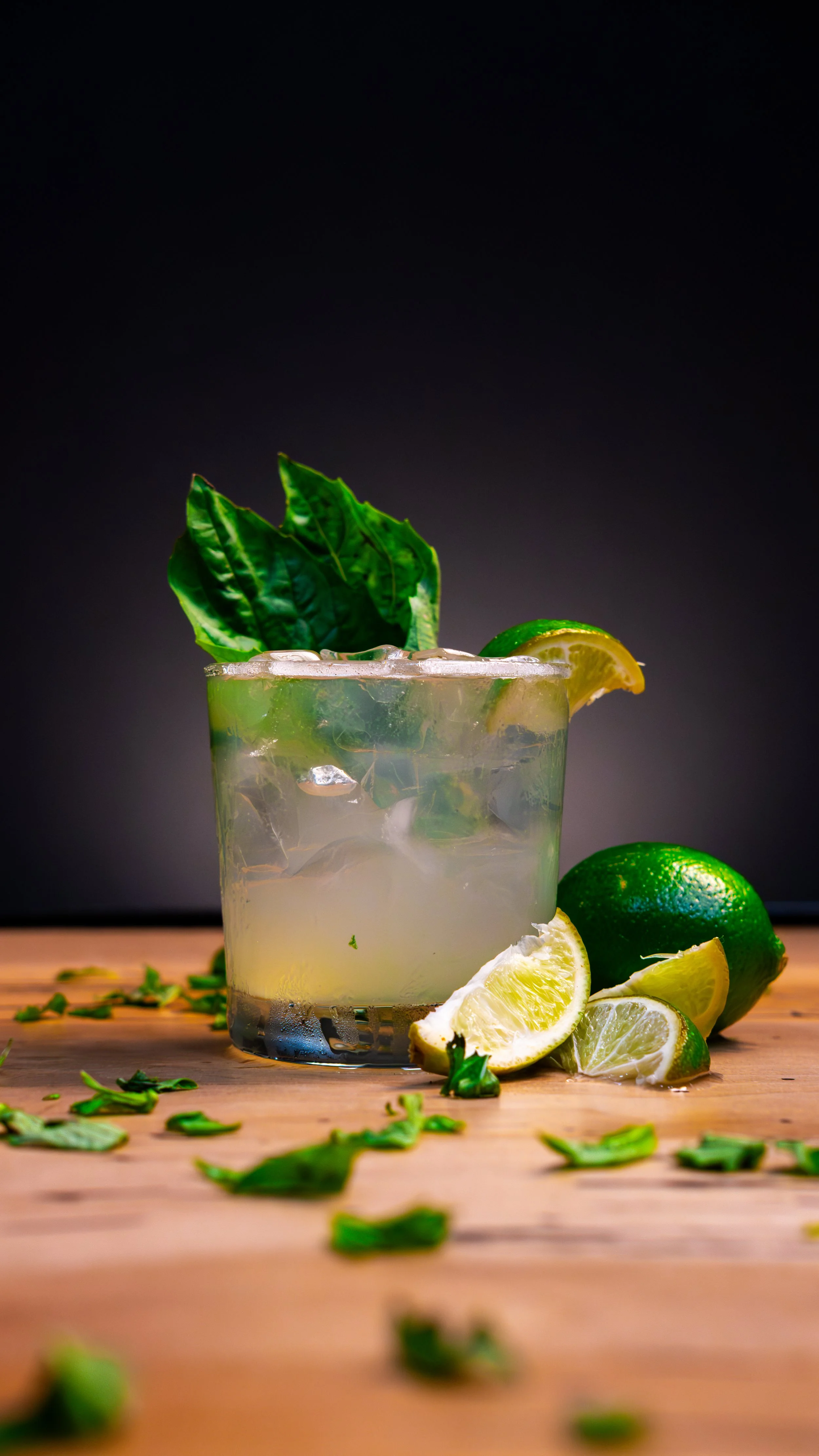 A glass of lime cocktail with ice, garnished with a lemon wedge, fresh basil leaves, and a whole lime on a wooden surface, with scattered basil leaves around.