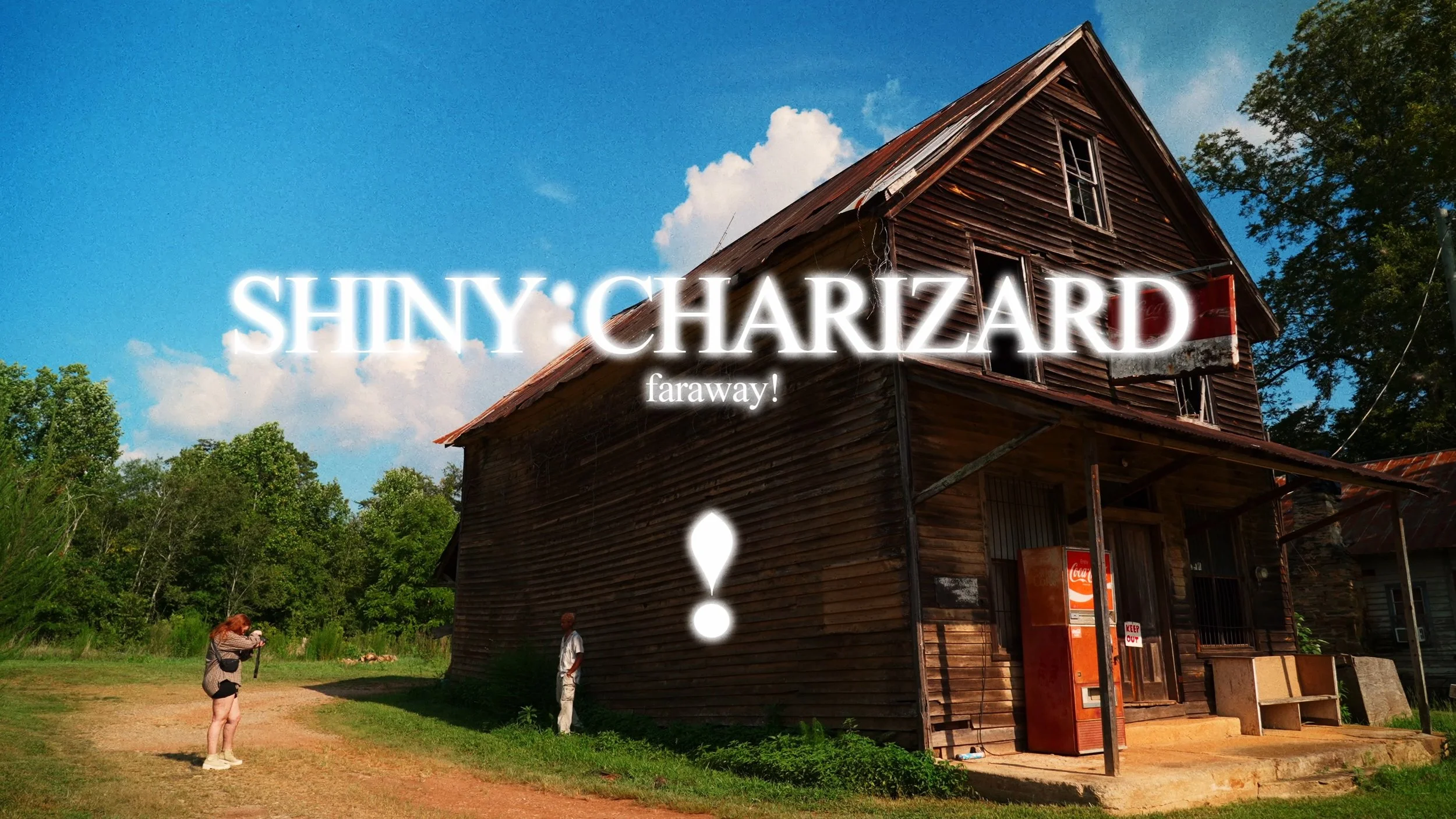 An old wooden house with a steep gabled roof in a rural area, surrounded by trees and blue sky with clouds. There are two people standing outside the house, one taking photographs and the other looking at them. The image has overlaid text that reads, 'SHINY: CHARZARD faraway!'