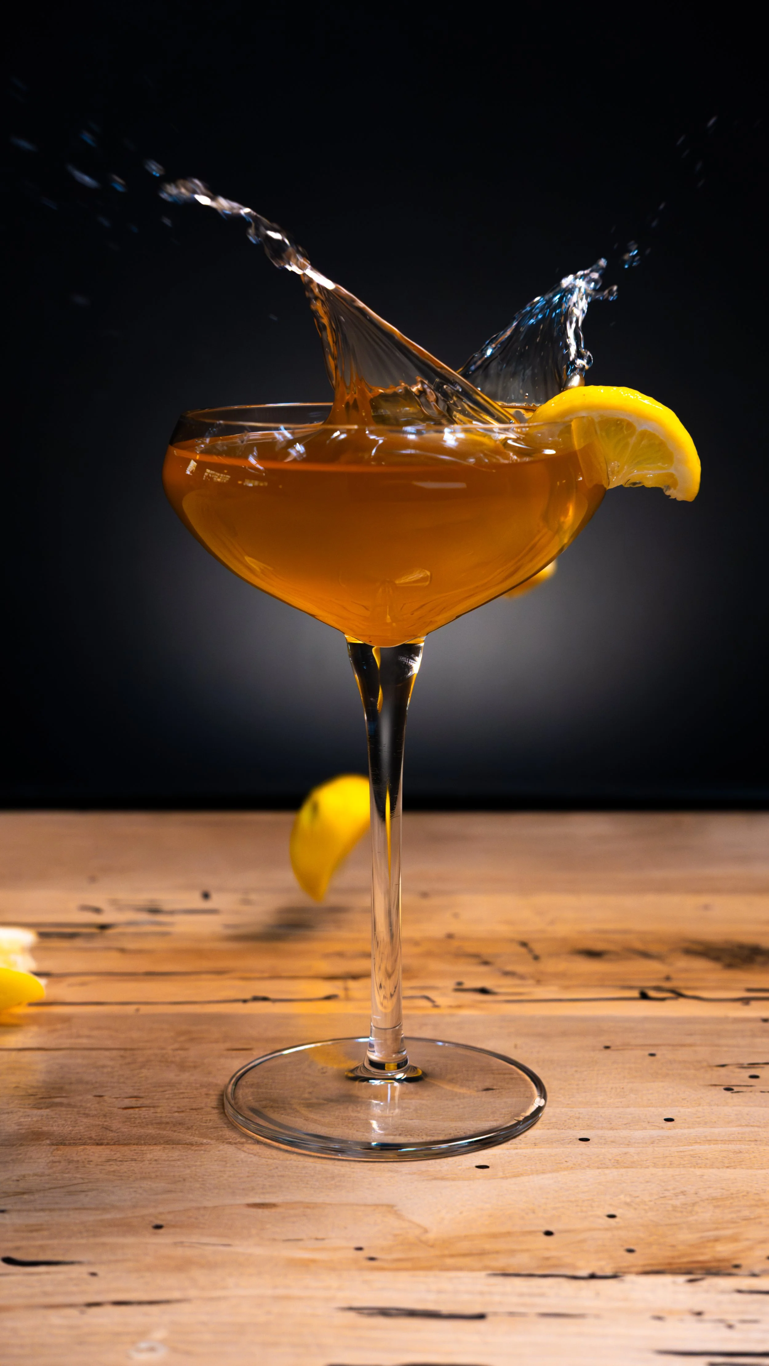 A cocktail glass filled with a dark orange beverage, garnished with lemon slices, with liquid splashing inside as it is poured, placed on a rustic wooden surface against a dark background.