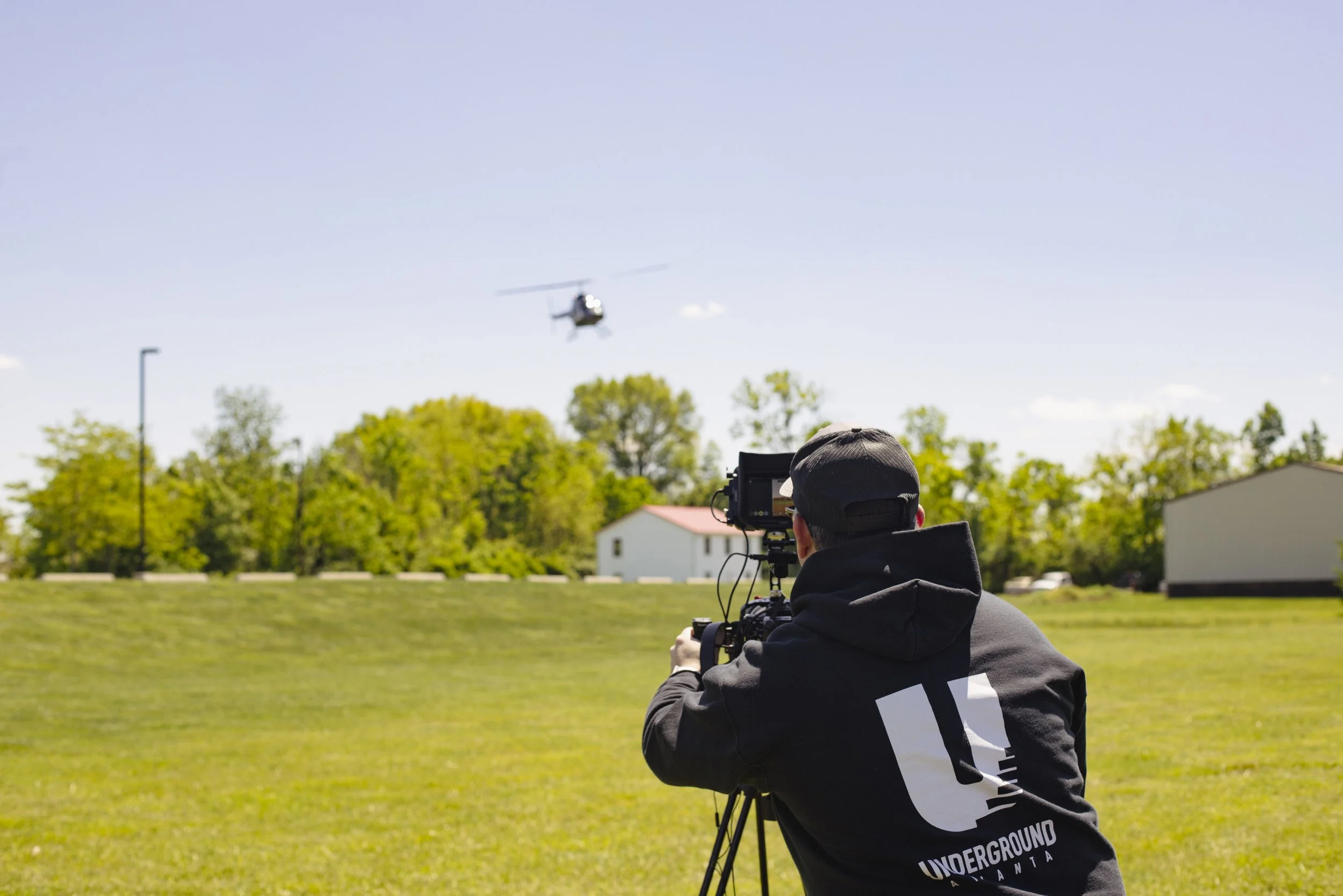 A person wearing a black hoodie and a cap is operating a camera on a tripod outdoors. A helicopter is flying in the sky in the background, with trees and buildings below.