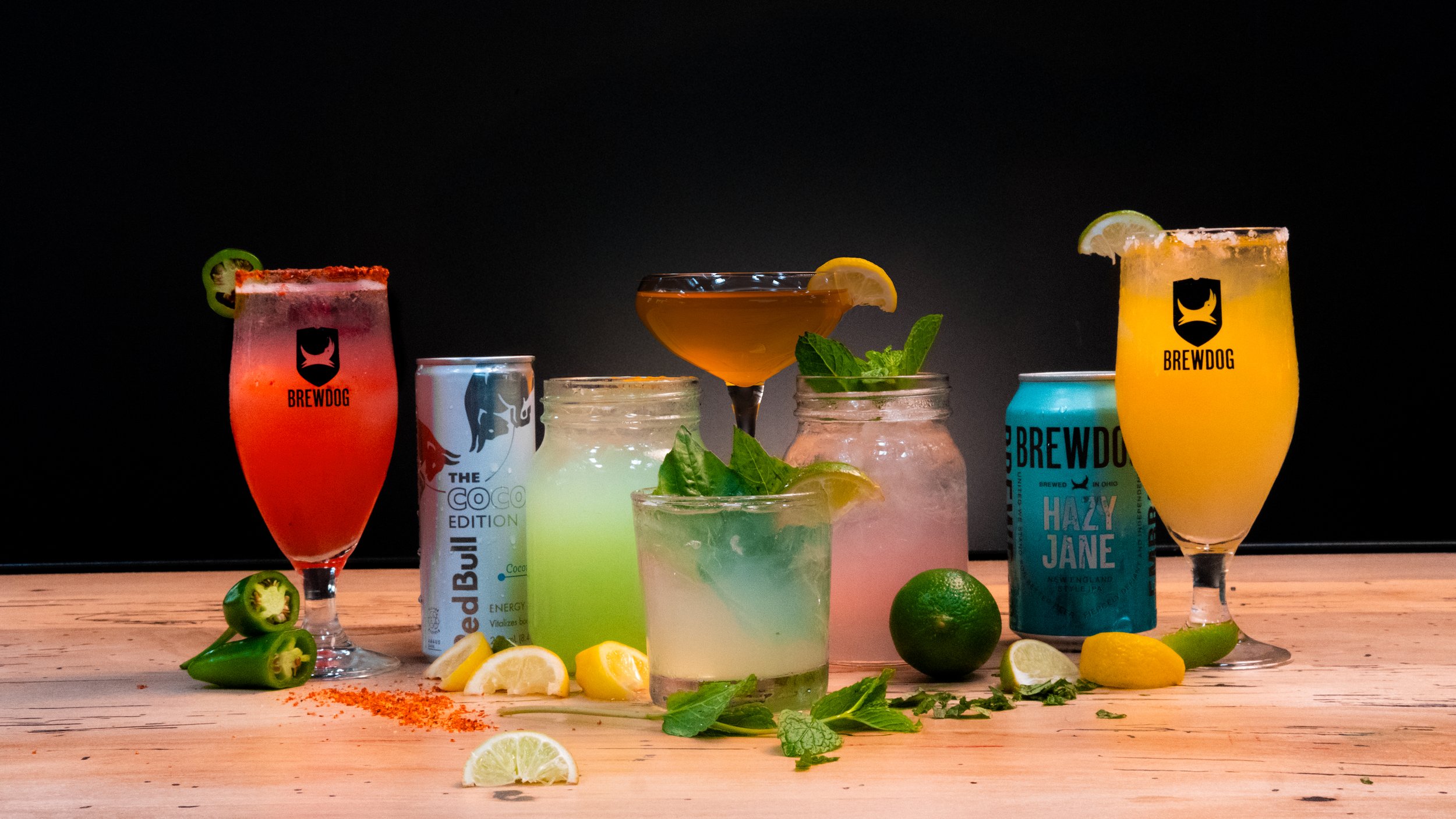 A variety of colorful cocktails with garnishes on a wooden surface, surrounded by lime wedges, lemon slices, mint, and pepper slices, with a black background.