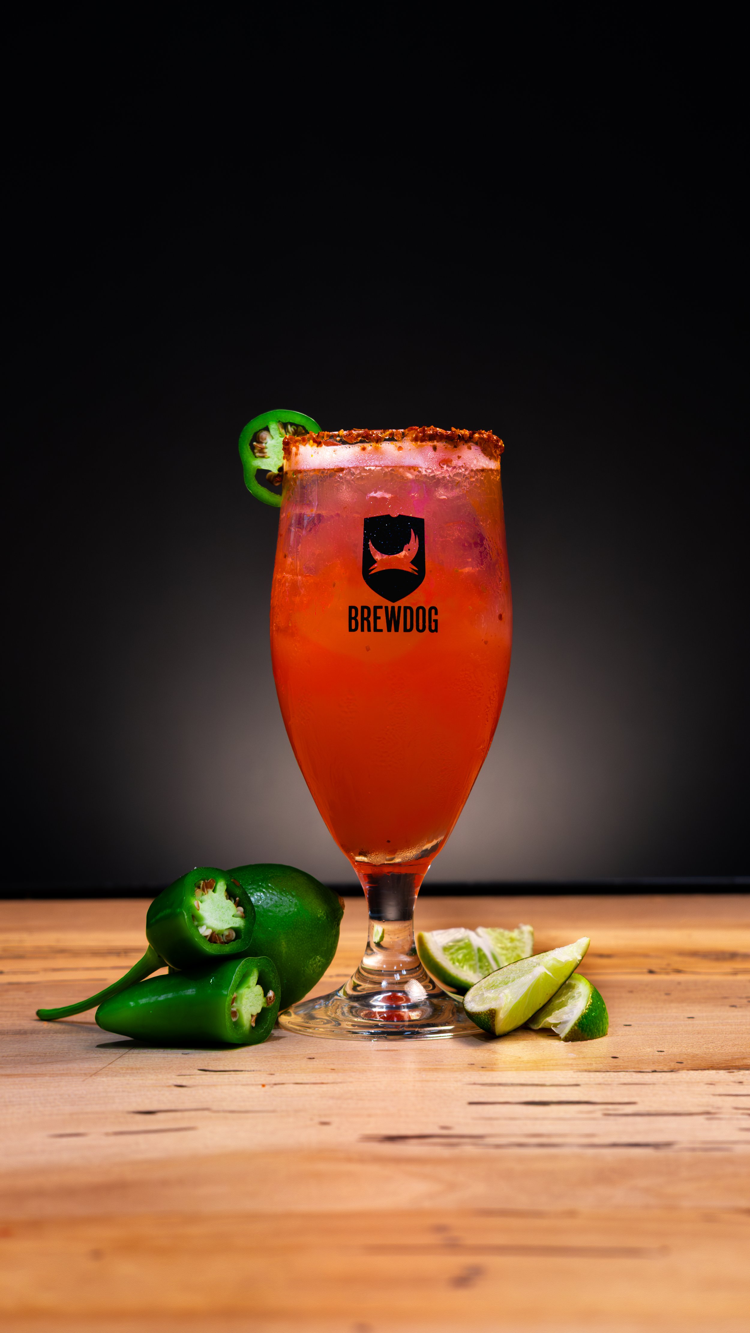 A festive pink cocktail in a tall glass with a salted rim, garnished with a slice of green jalapeño, on a wooden surface with sliced limes and whole jalapeños nearby, branded with the Brewdog logo.