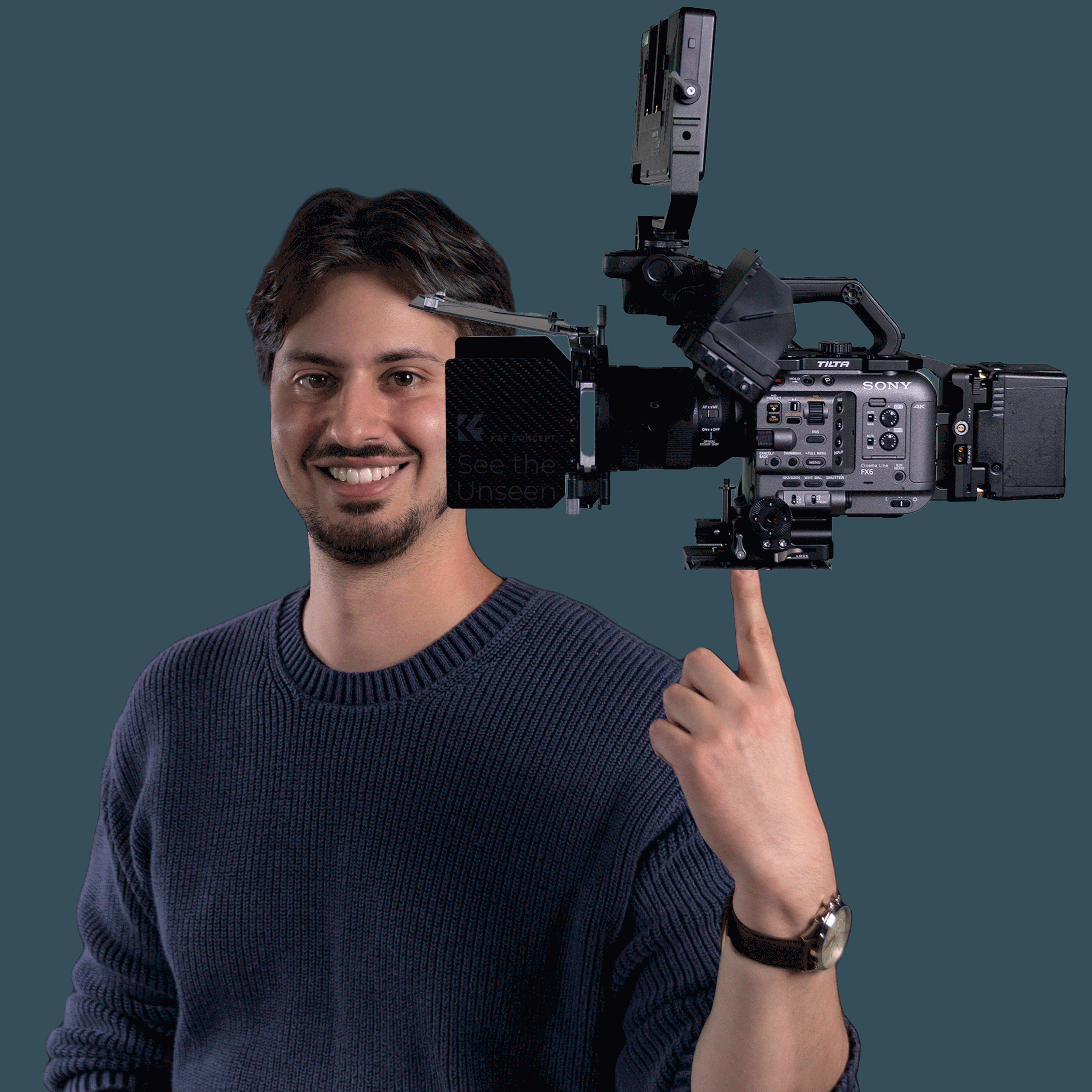 A man with brown hair and a beard smiling as he holds a professional video camera on his shoulder, wearing a dark sweater and a wristwatch, against a plain dark background.