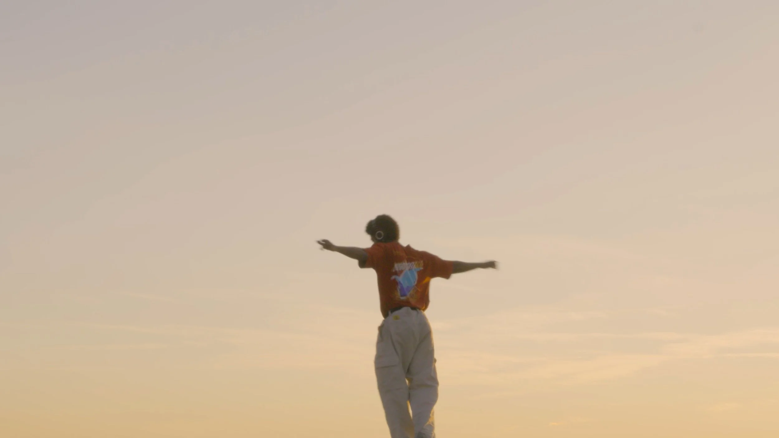 A person balancing on another person's shoulders with arms outstretched, against a sunset sky.