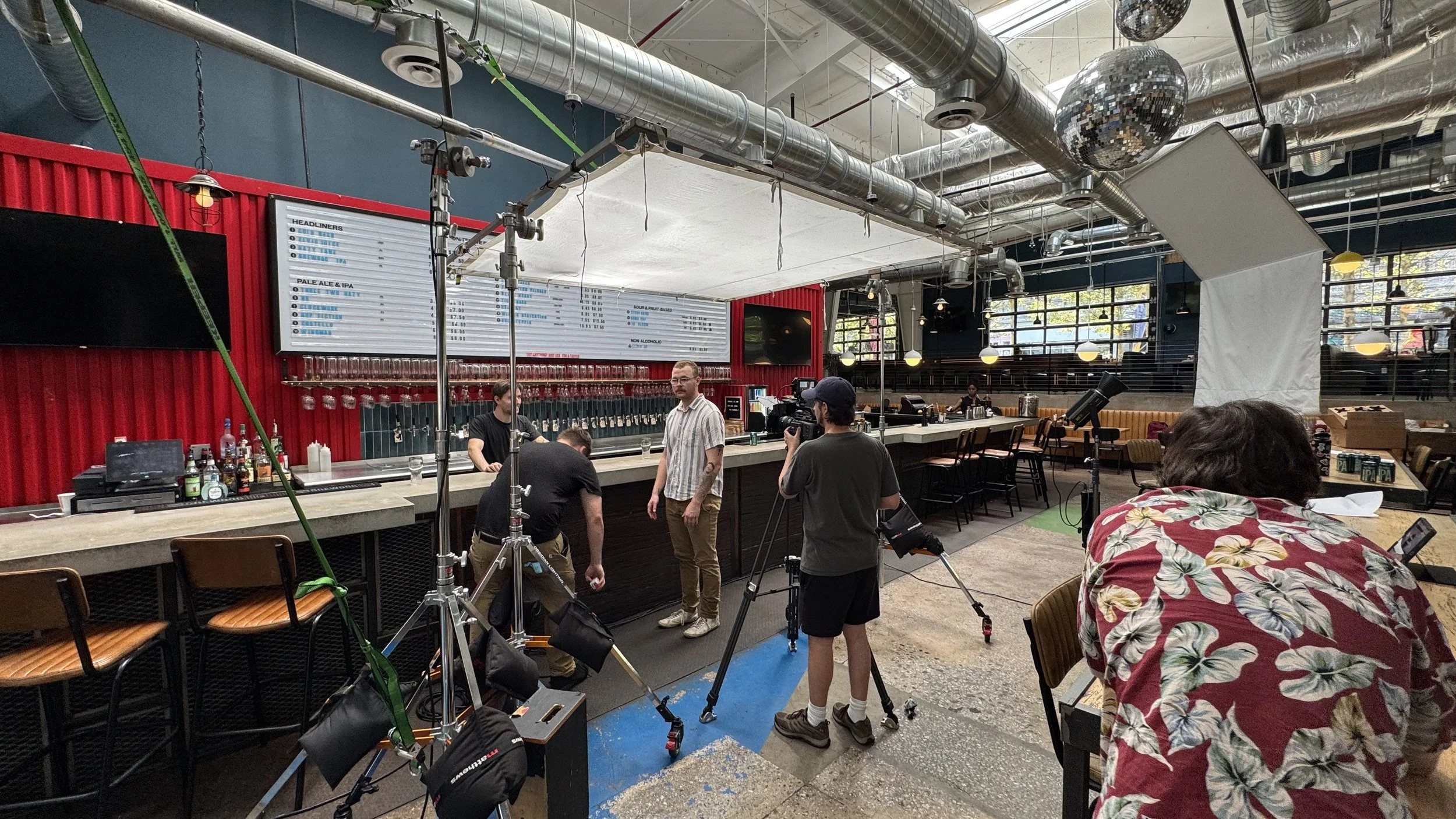 Filming crew setting up equipment inside a bar or restaurant with a red wall, large menu boards, and large windows, preparing for a shot or interview.