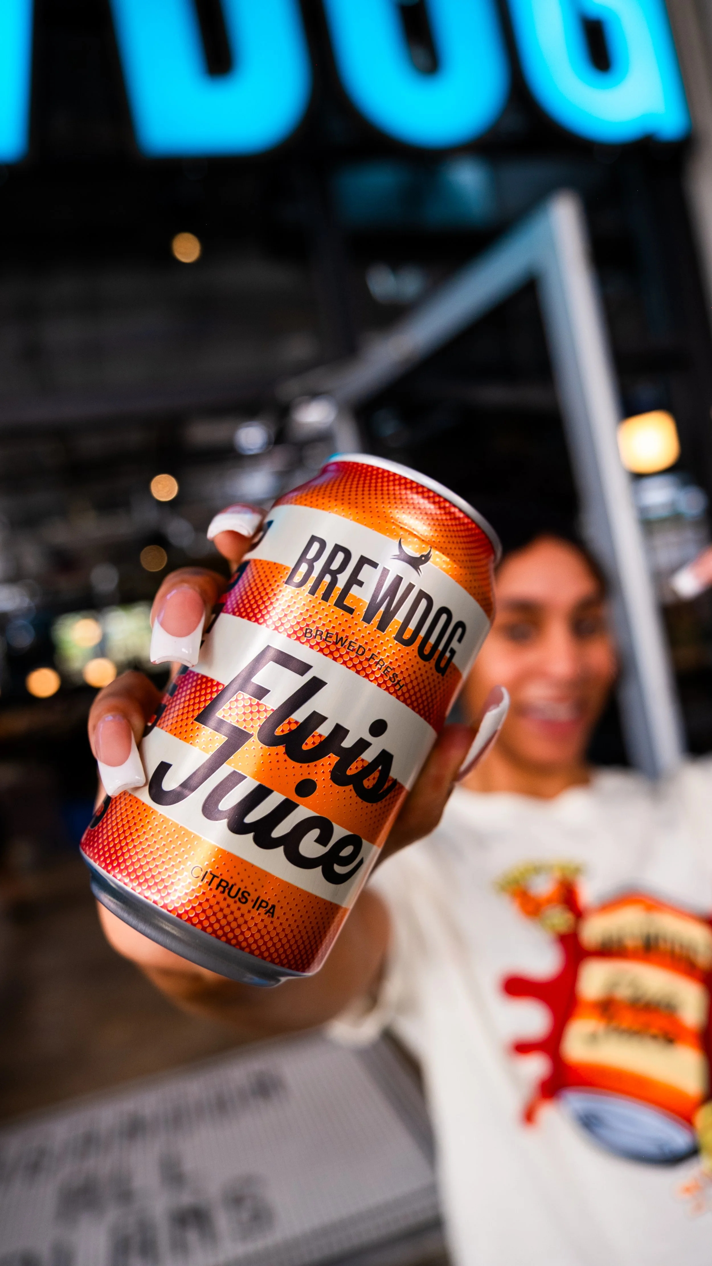 A woman holding a can of Brewdog Citrus IPA beer in front of her, with a blurry background featuring a woman smiling and a neon sign.
