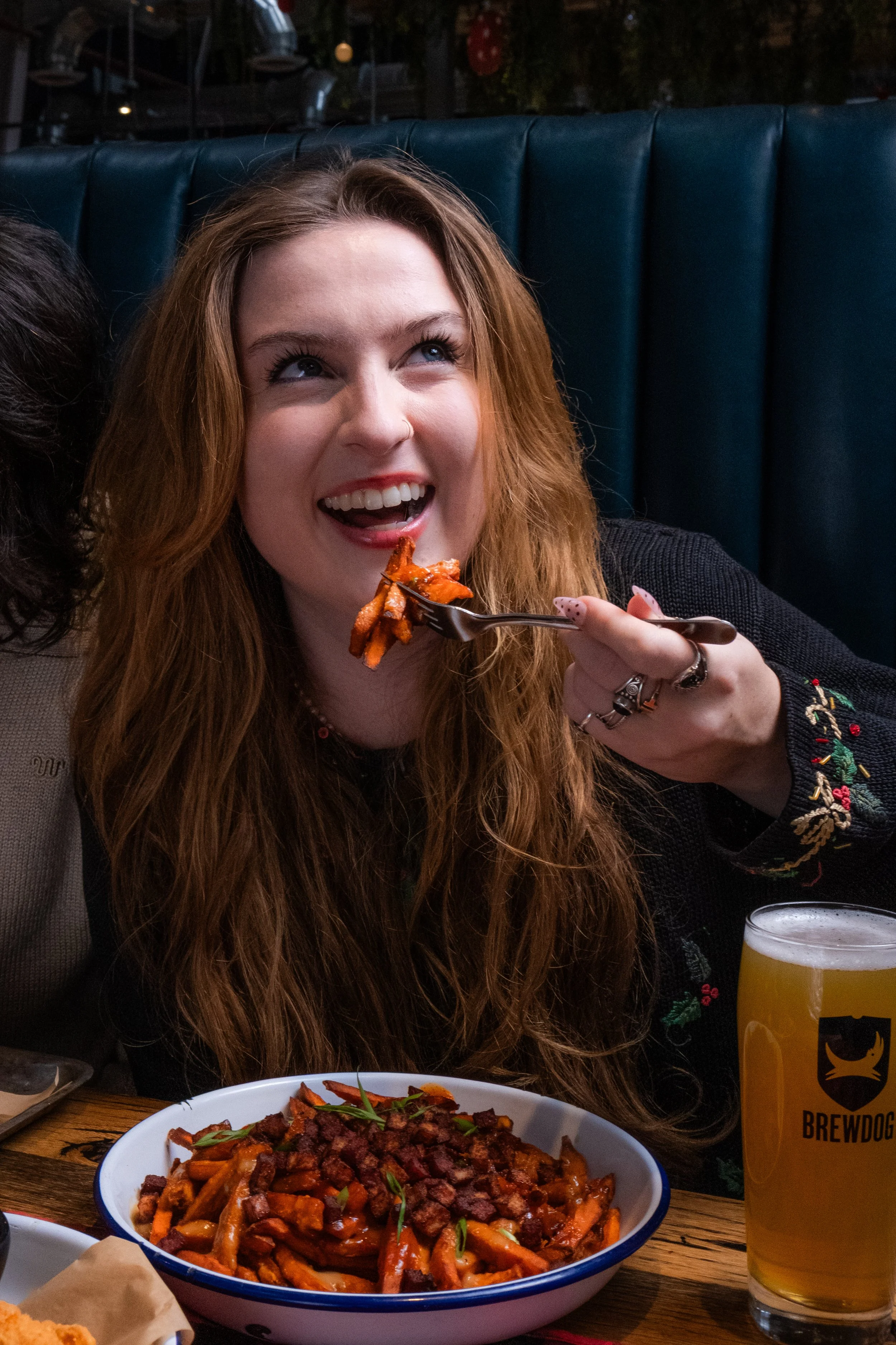 A woman with red hair and a nose ring sitting at a table in a restaurant, eating pasta. There is a large bowl of pasta with sauce and chopped meat, and a tall glass of beer with a Brewdog logo on it. The background shows a booth with dark green uphol