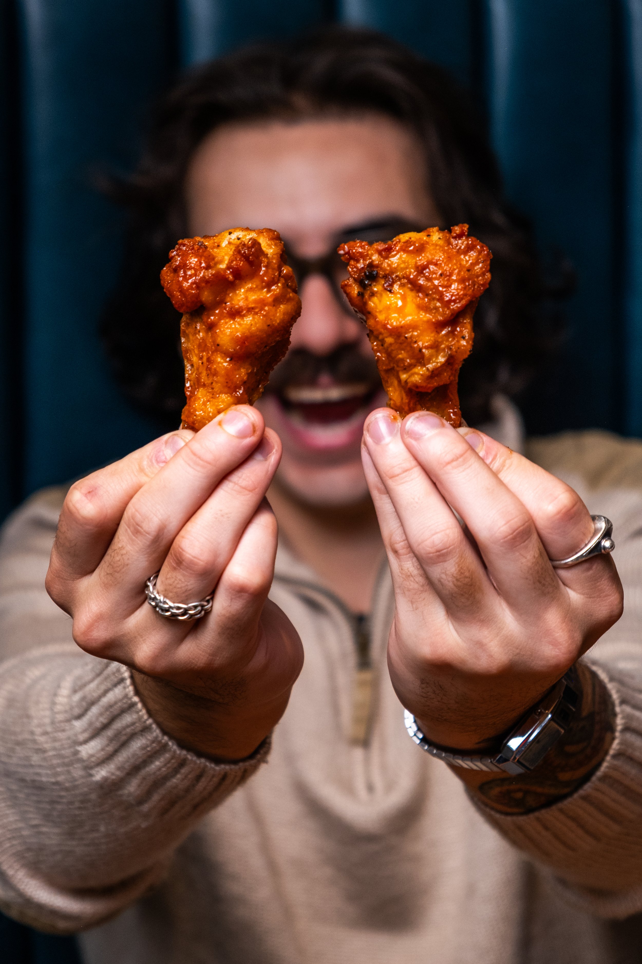 Person holding two fried chicken drumsticks in front of their face, with a dark blue background.