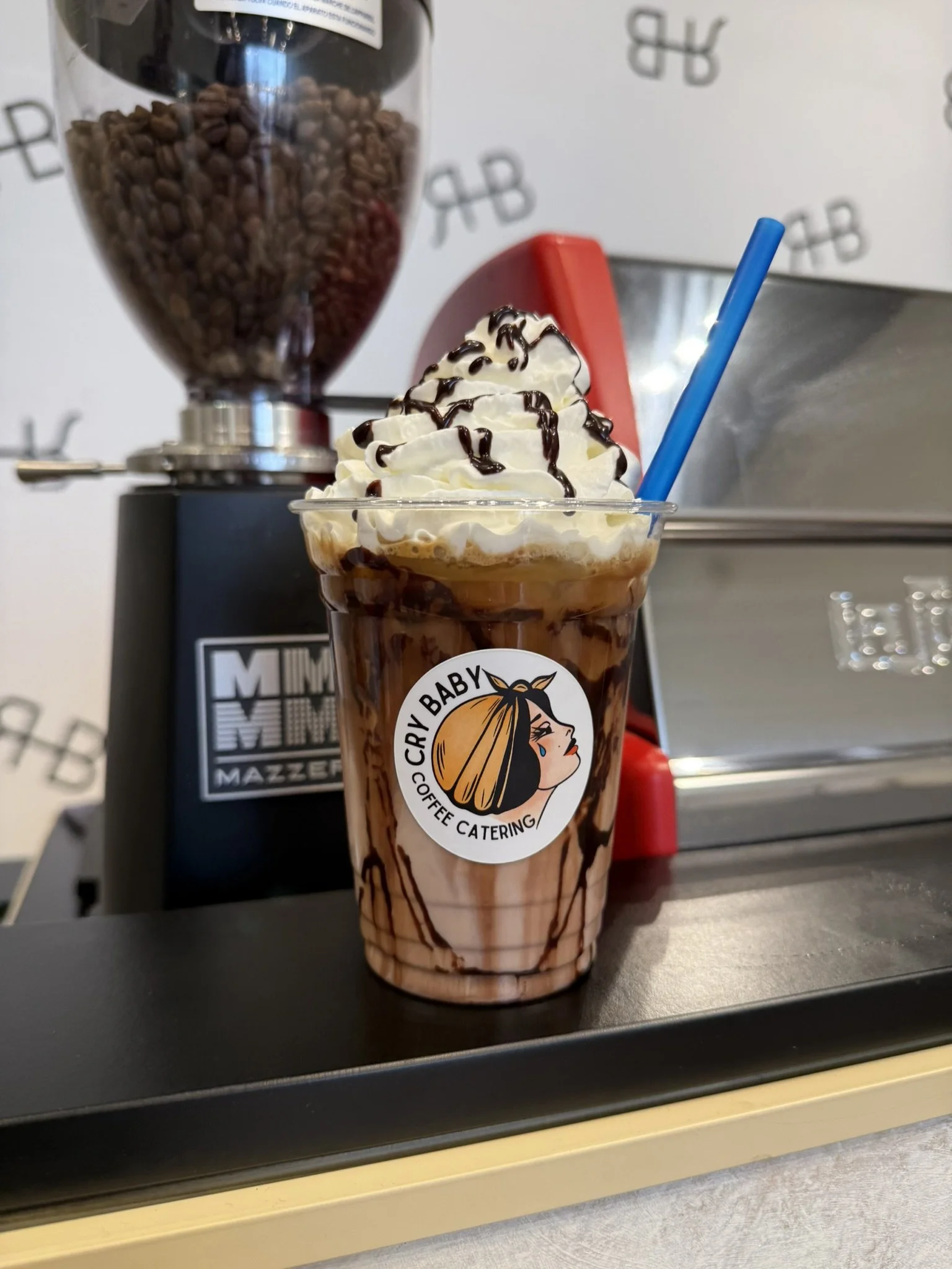 Iced mocha with whipped cream and chocolate drizzle in a Cry Baby Coffee Catering cup on espresso machine.