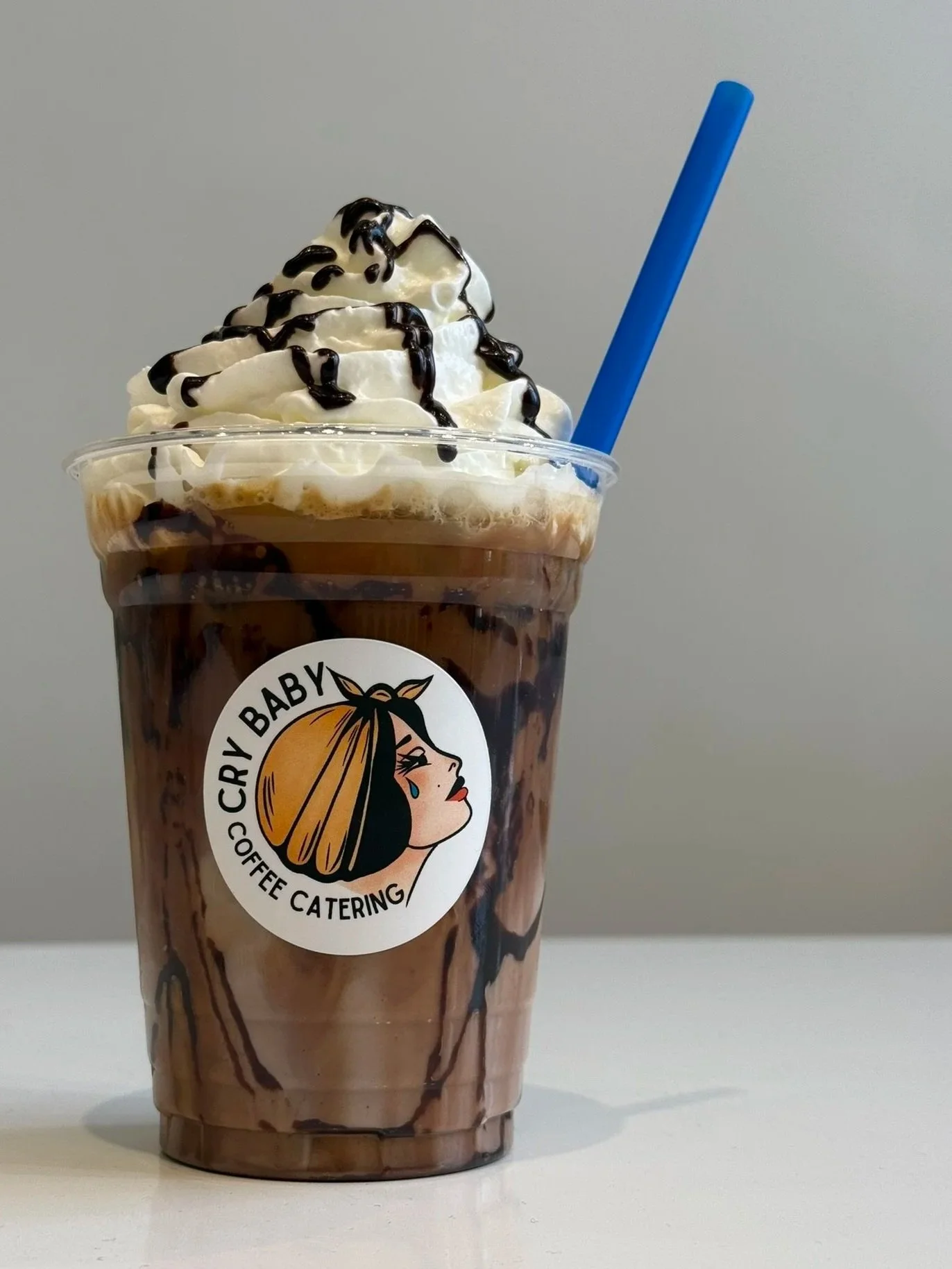 Iced mocha with whipped cream and chocolate drizzle in a branded Cry Baby Coffee cup with a blue straw