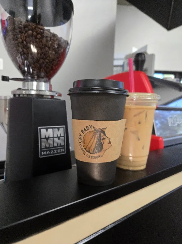 Cry Baby Coffee Catering hot coffee cup with logo and iced latte next to espresso grinder with coffee beans.