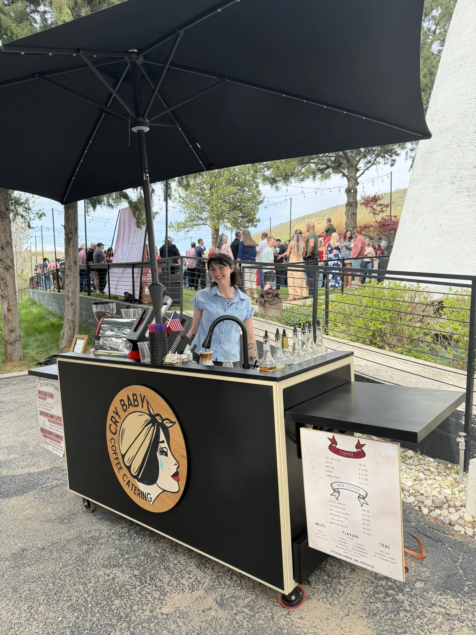Cry Baby Coffee Catering espresso cart set up outdoors under an umbrella, with a barista serving drinks from a branded mobile coffee bar while guests gather at a social event in the background.