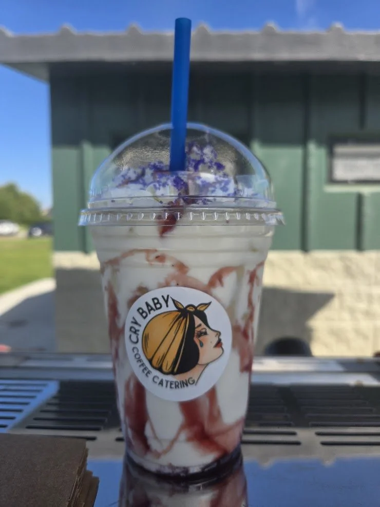 Iced drink with cream, berry drizzle, and whipped topping in a Cry Baby Coffee Catering cup outdoors.