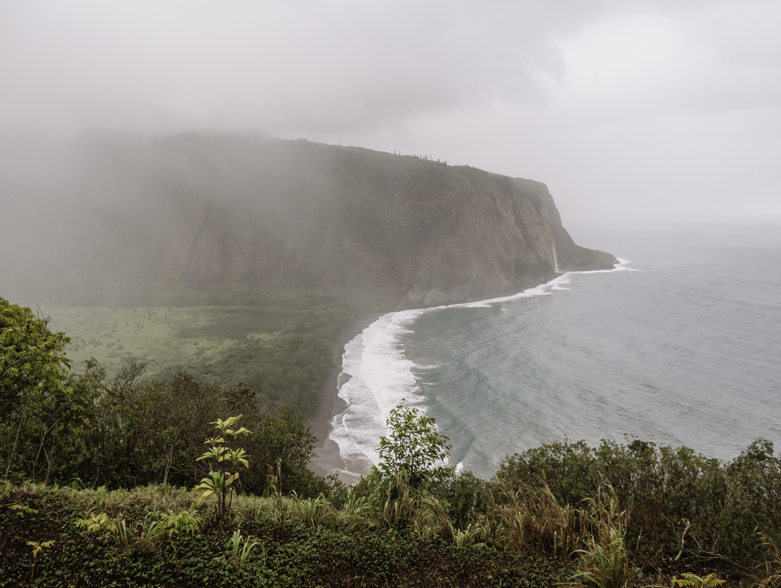 Misty view of Waipiʻo Valley in Hawaiʻi with steep green cliffs, a black sand beach, ocean waves, and a distant waterfall cascading into the shoreline.
