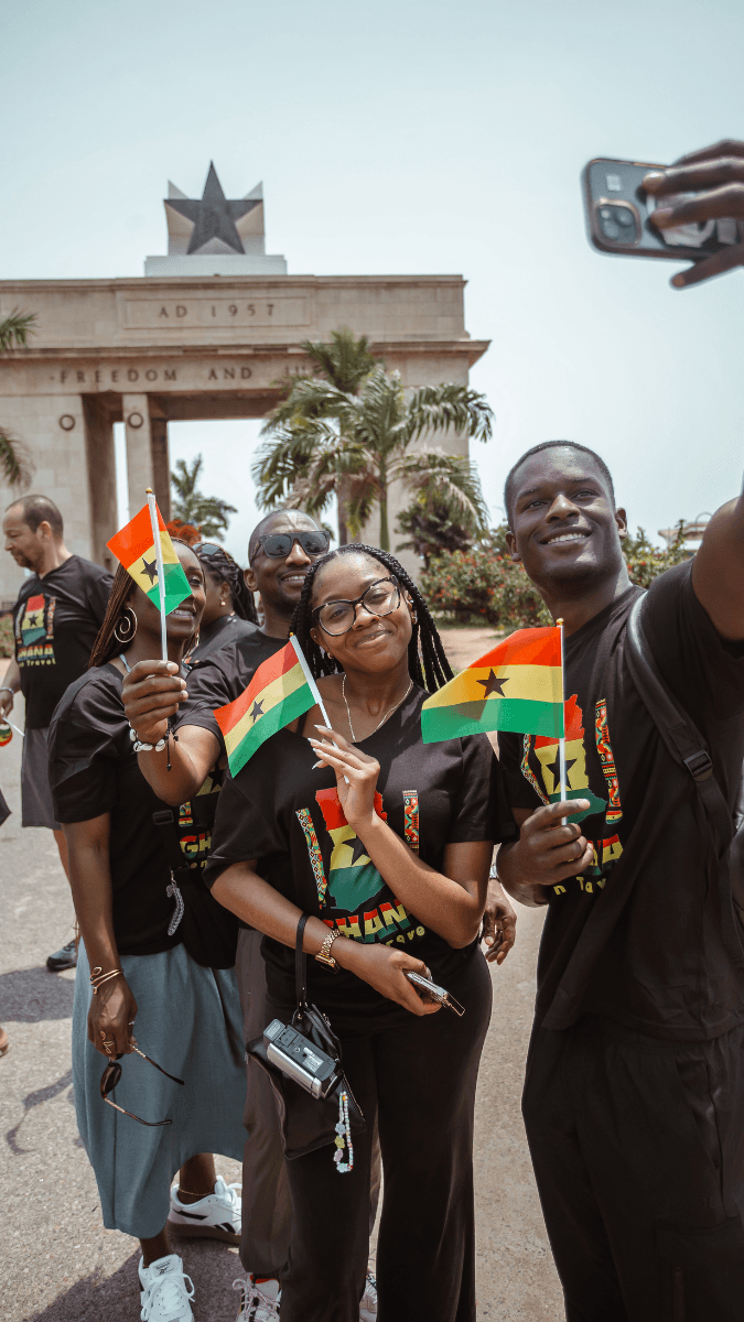 A family of four taking a photo in front of the Black Star Square monument holding Ghanaian flags, wearing black T-shirts with Ghanaian colors.