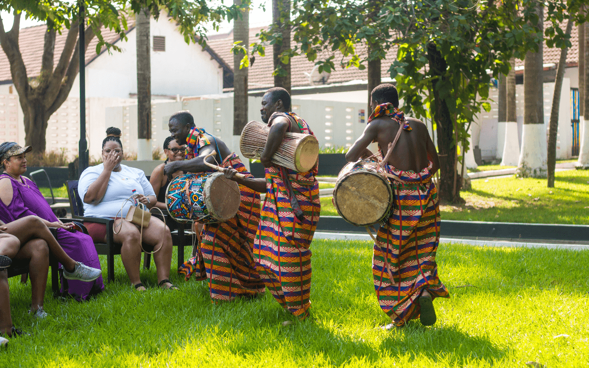 Three men in colorful patterned traditional clothing playing drums and percussion instruments while singing outdoors during a drumming and dance tour experience