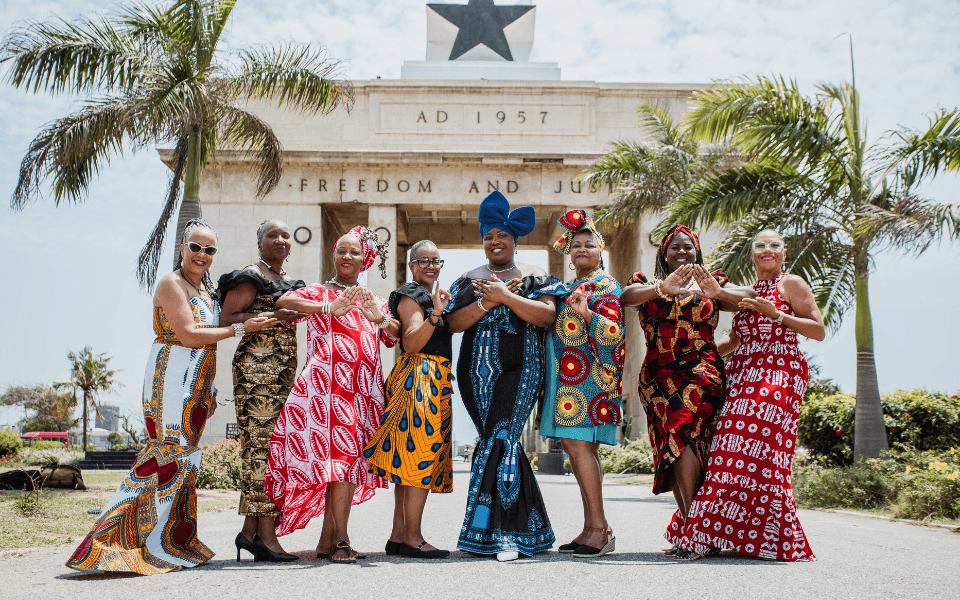 A group of women dressed in African Attire standing in front of the Black star square monument in Ghana