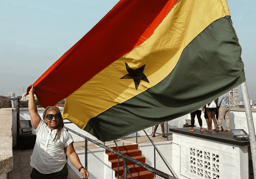 A woman wearing sunglasses and a white shirt is holding a large Ghanaian flag on a rooftop with other people in the background.