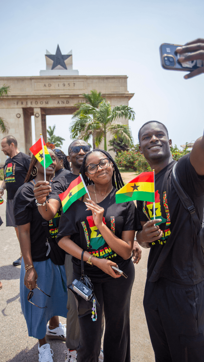 A family of four taking a photo in front of the Black Star Square monument holding Ghanaian flags, wearing black T-shirts with Ghanaian colors.