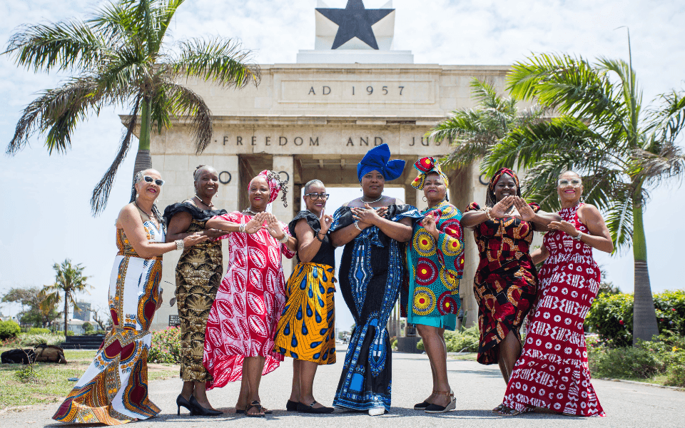 A group of diaspora women dressed in African print  dresses posing for a photo in front of the Black Star Square monument in Ghana
