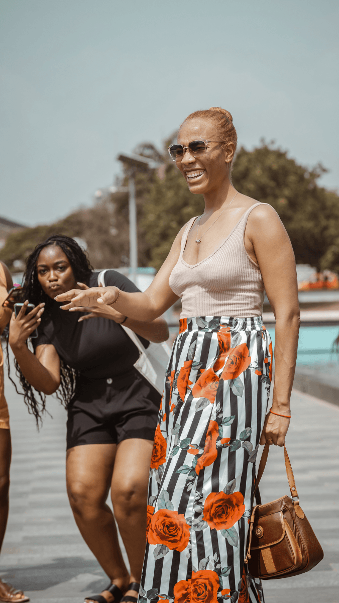 A woman in sunglasses, a beige tank top, and a floral skirt with red roses and black and white stripes, smiling and talking at the Kwame Nkrumah Memorial Park