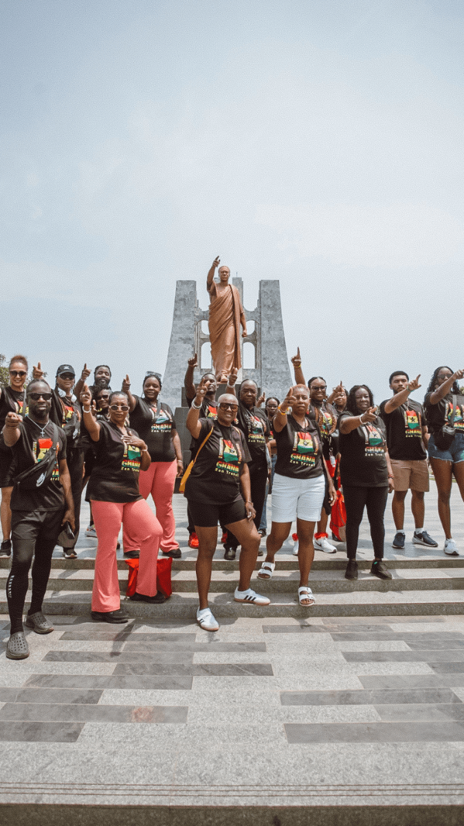 A group of tourists standing in front of the Kwame Nkrumah se of a man in orange robes, with many raising their index fingers